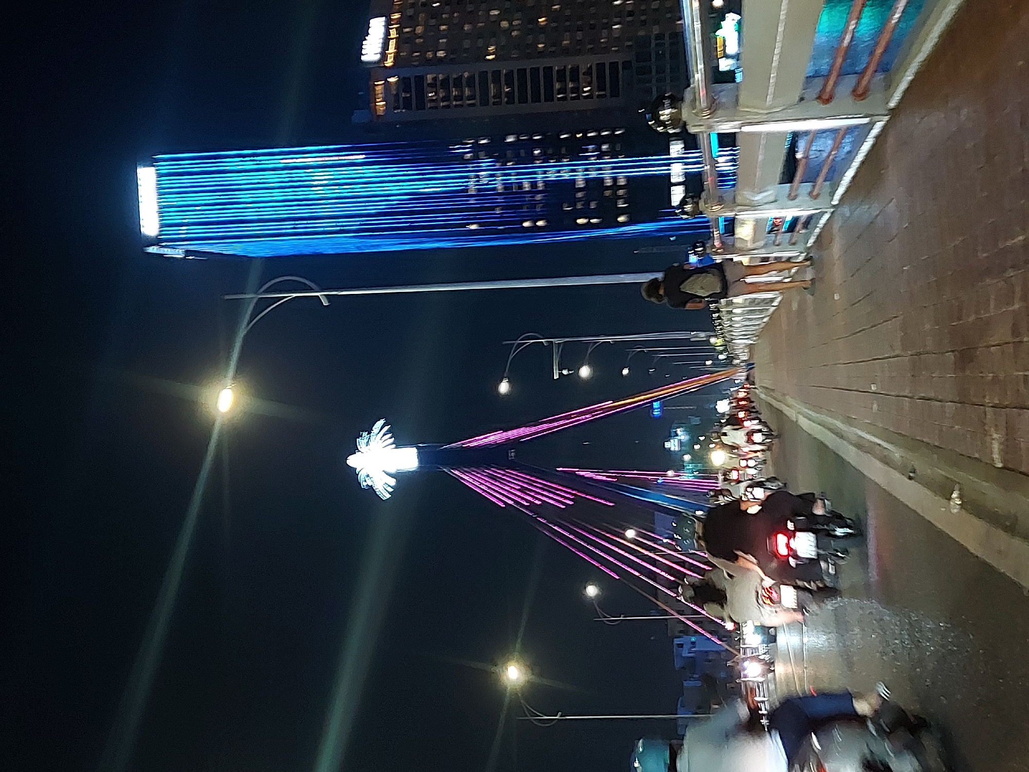 This is a night-time, vertical, street-level shot taken from a pedestrian walkway on a bridge in Da Nang, Vietnam. The bridge is illuminated with pink and purple lights on its suspension cables, leading to a prominent, brightly lit, stylized lotus flower structure at its apex. To the right, a tall, modern skyscraper is covered in vertical blue lights. A Hilton hotel sign is visible on another building to the right of the skyscraper. The foreground shows a pedestrian walkway paved with brick, and to the left, the bridge's roadway is filled with motorbikes in motion, their headlights and taillights creating streaks of light. One person is walking on the pedestrian walkway, facing away from the camera. The scene is bustling with activity despite the late hour, illuminated by a combination of the bridge's decorative lighting, the skyscraper's modern facade, and vehicle lights.