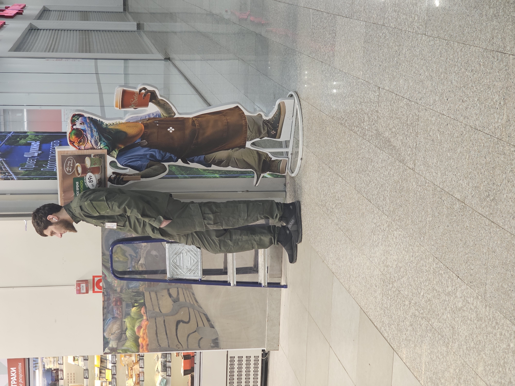 An indoor scene is depicted within a commercial hallway in the Shchukino district of Moscow, Russia. The hallway features light-colored walls, a reflective speckled floor, and overhead artificial lighting.
On the left, a Caucasian male adult with dark hair, a beard, and a mustache stands, leaning slightly forward with his hands in his pockets. He wears an olive green utility-style uniform with an ID badge clipped to his chest, black Adidas sneakers, and a white in-ear headphone in his right ear. He faces right, away from the camera.
Positioned to his right is a blue metal stepladder, partially folded and resting against a large reflective panel that displays an image of various fruits and a large bird. Further to the right stands a life-sized cardboard cutout of an anthropomorphic lizard or iguana. This cutout wears a blue shirt, a brown apron, sunglasses, and a colorful headscarf, holding a brown coffee cup labeled "Select" and advertising coffee priced from 89 rubles.
In the background, to the far left, a refrigerated display case containing various food products is visible beneath a sign with partially visible Russian text reading "ТРАКИ" and "е рецепты." A large banner behind the cutout displays Russian text, including "Привет, Щукино!" (Hello, Shchukino!) and "Добро пожаловать." The right side of the hallway shows multiple storefronts with metallic roller shutters, indicating they are closed.