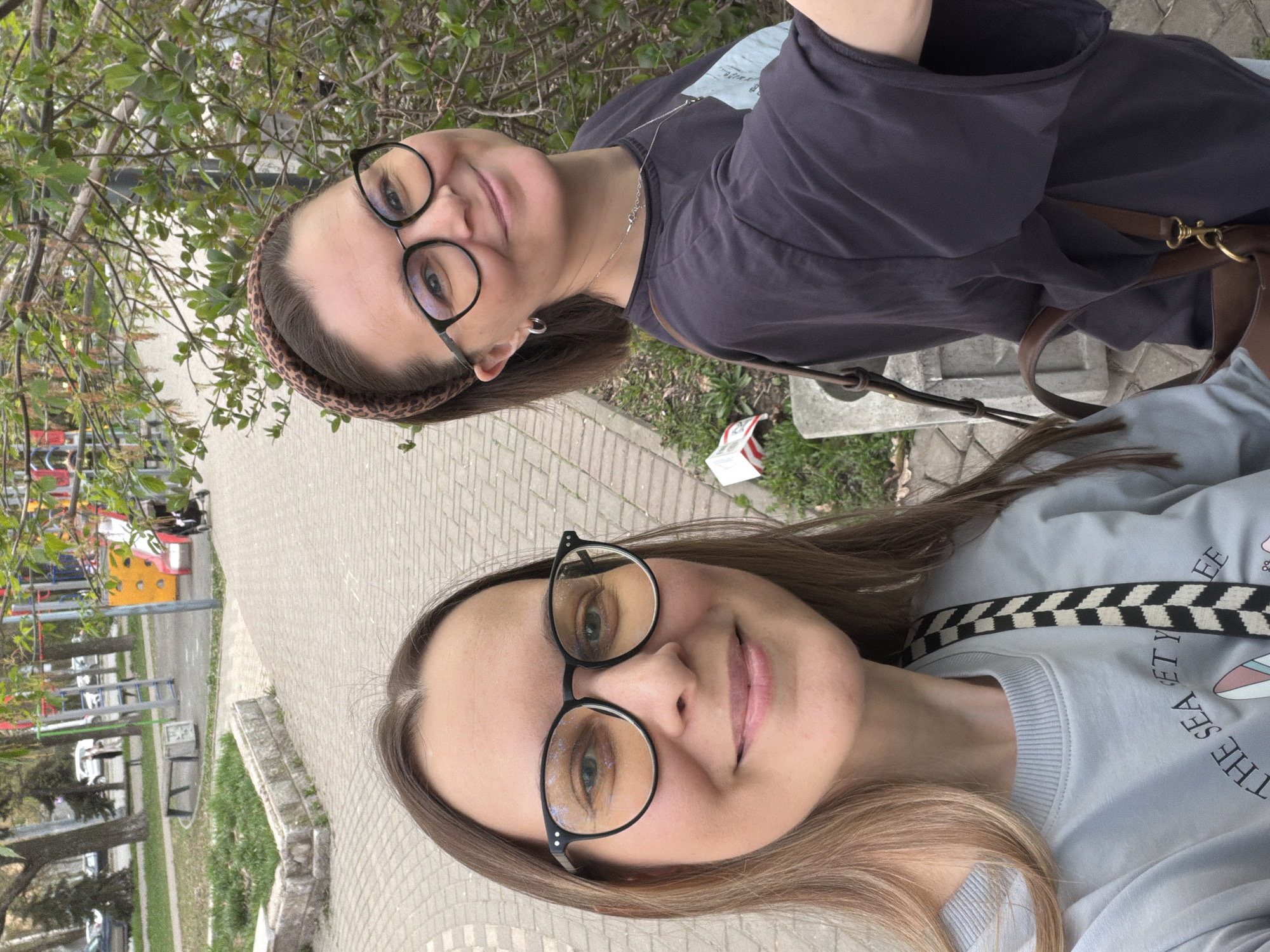 Two women are posing outdoors on a paved surface. The woman in the foreground wears eyeglasses, a light blue sweater with a checkered strap across her shoulder. The woman in the background also wears eyeglasses and a patterned headband. She is wearing a dark-colored shirt. They appear to be in an urban environment, with trees and a playground visible in the background. A brick walkway is visible. Based on the background details, the location is likely in Izhevsk, Russia.