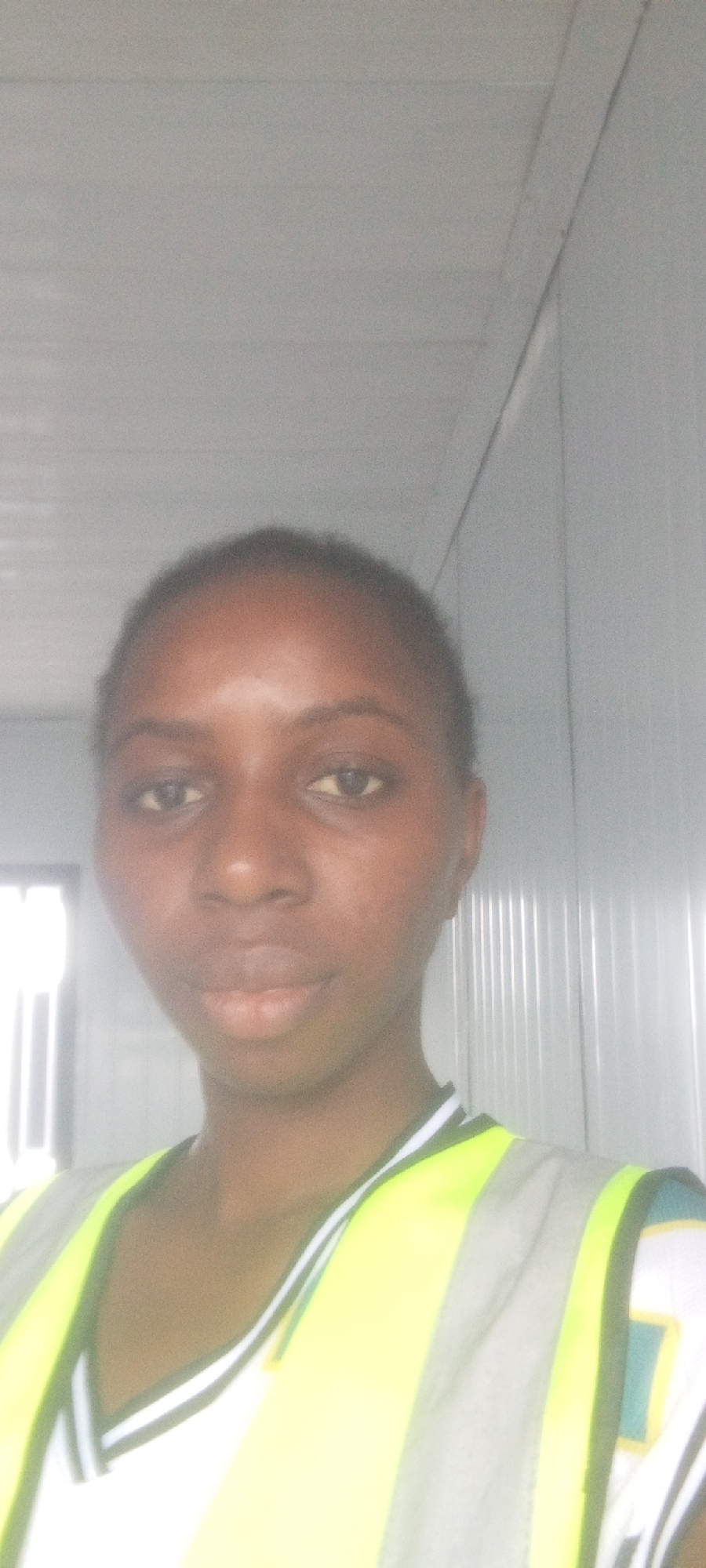 A medium shot selfie shows a woman with dark skin and dark hair. She is wearing a white t-shirt and a high-visibility vest with reflective strips. The vest is lime green and gray. The background appears to be an indoor structure with ribbed metal walls and a white ceiling, possibly in Indo, Nigeria. The lighting is bright and somewhat washed out. The woman is looking directly at the camera with a neutral expression.
