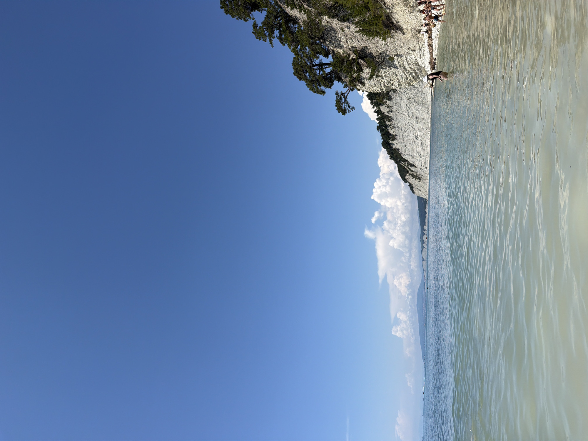 The image captures a view of the sea from a shallow beach on a clear, sunny day. In the foreground, gentle waves lap at the shore, with the water appearing clear and shallow. The background features a dramatic, white rocky cliff face covered with lush green trees, characteristic of the Divnomorskoe, Russia coastline. A thin strip of beach is visible along the base of the cliff, with a few people present. Further in the distance, the sea stretches out to the horizon, meeting a hazy sky. A bridge and distant landmasses are faintly visible on the left side of the frame. The overall scene depicts a serene coastal environment.