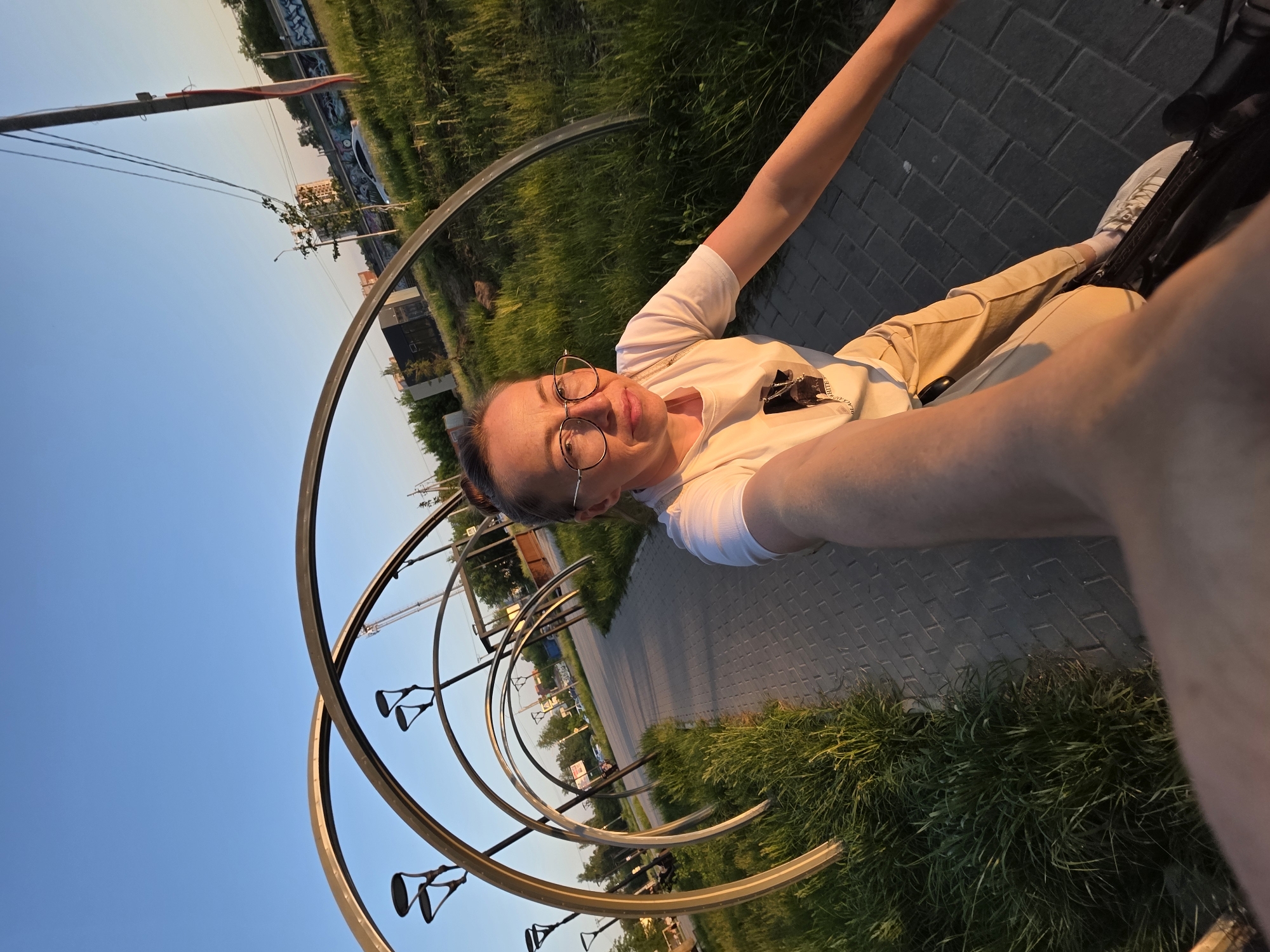 A woman wearing a white t-shirt and beige pants stands in front of an art installation in Chaikovsky, Russia. The installation consists of a series of concentric metal rings supported by vertical posts. The rings are located on a paved path next to a grassy area. A building with a mural is visible in the background. The sky is blue. The woman is holding the camera up to take a selfie.