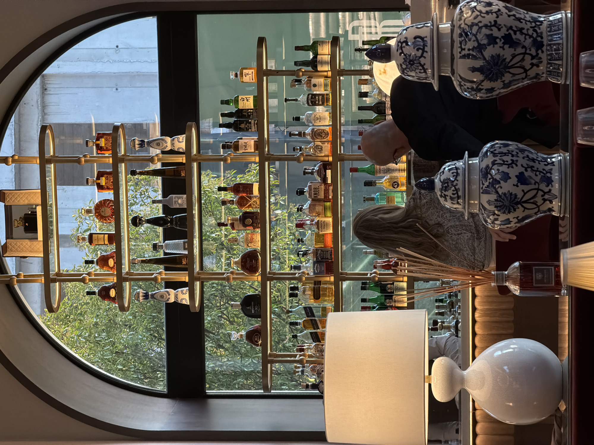 The image captures the interior of a bar or restaurant in Belgrade, Serbia. Prominently featured is a window with a unique rounded arch, offering a view of a concrete building and some greenery. A gold-colored metal rack with several shelves filled with various bottles of liquor stands against the window, seemingly functioning as a decorative display. In the foreground, a table with a white lamp and two blue and white porcelain vases partially obscures the view. Two individuals are visible near the rack, one appearing to be a bald man examining the bottles, and the other with long hair. Diffusers are visible on the table near the lamp and vases.
