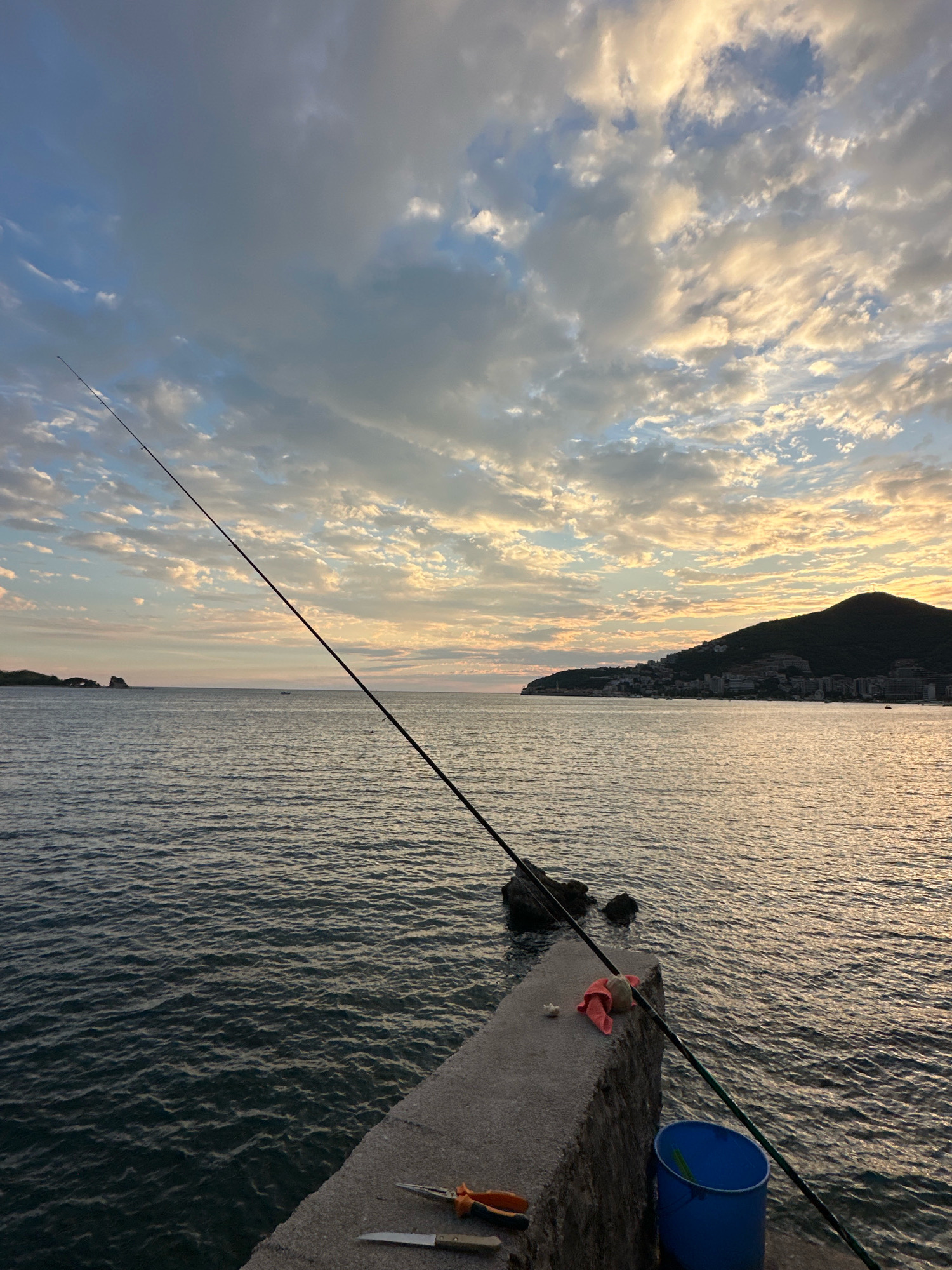 This image captures a serene coastal scene likely in or near Budva, Montenegro, at dusk. The foreground features a concrete pier where fishing equipment is laid out. A long fishing rod extends diagonally across the frame, its tip disappearing into the cloudy sky, which is illuminated by the warm hues of the setting sun. Near the fishing rod, on the pier, are a pair of pliers, a knife, and a pink cloth. A blue bucket is also visible. The sea, rendered in shades of deep blue and grey, stretches to the horizon, with gentle ripples on its surface. In the distance, a hilly coastline with buildings and a prominent mountain is silhouetted against the illuminated sky. A small island or rock formation is visible to the left in the sea. The overall atmosphere is one of peaceful anticipation, possibly of a successful fishing outing.