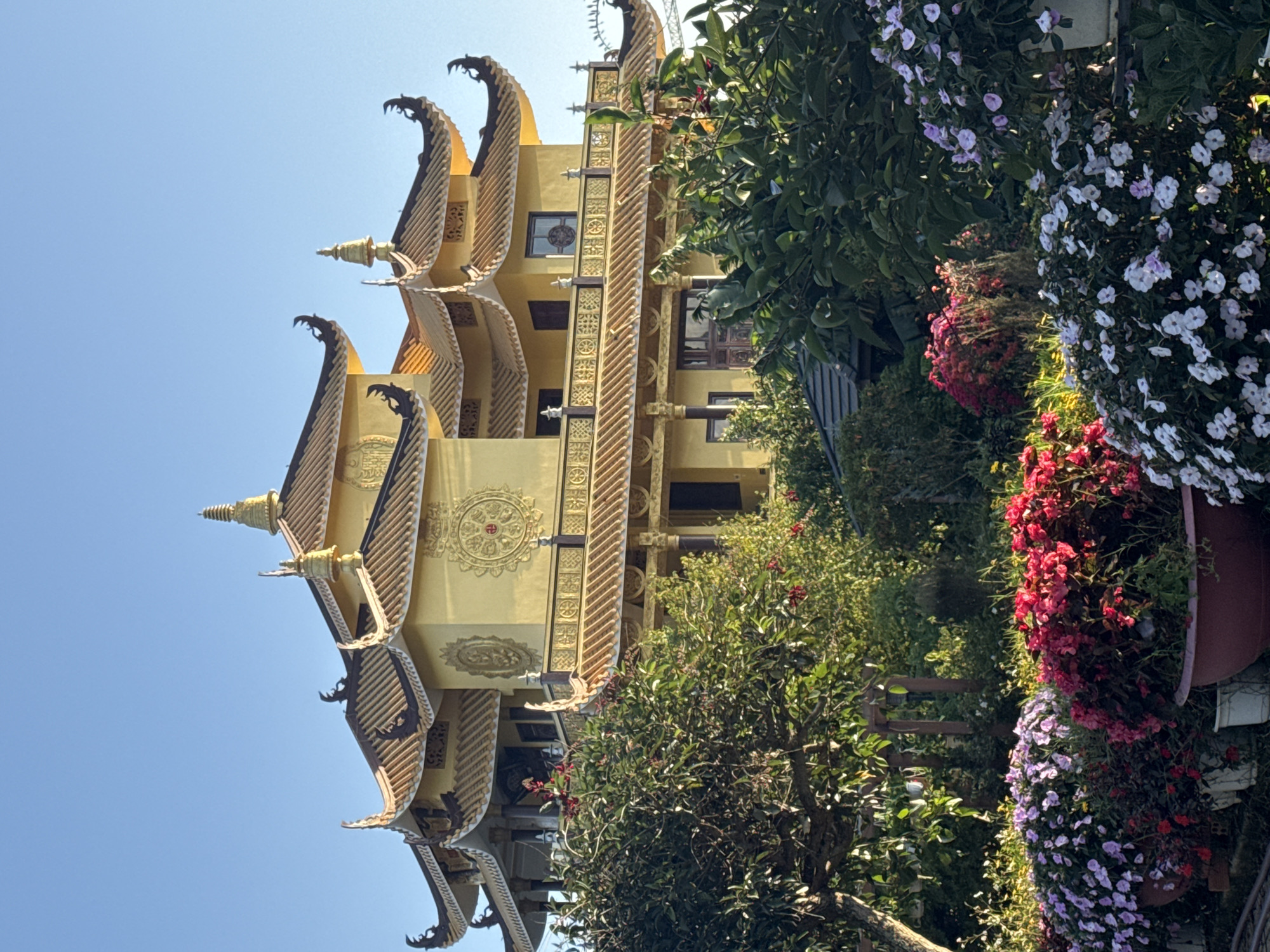 The image captures a vibrant Buddhist temple with traditional yellow architecture set against a clear blue sky. The temple's multi-tiered roofs are adorned with intricate golden decorations and finials. Lush greenery, including trees and colorful flower beds filled with pink, red, and purple blossoms, surrounds the temple, creating a picturesque foreground. The scene appears to be an outdoor setting, possibly a garden or temple grounds, bathed in natural sunlight. This specific temple is likely Truc Lam Zen Monastery, a prominent Buddhist monastery located on Phung Hoang Mountain in Da Lat, Vietnam.