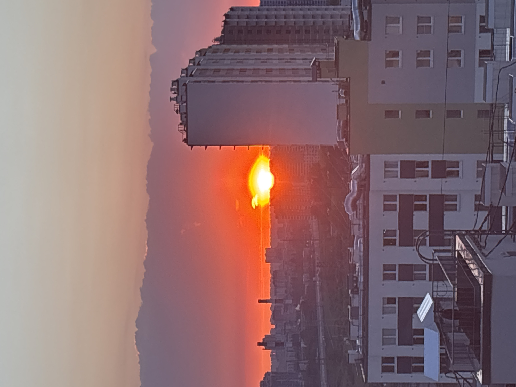 The image shows an urban sunset in Vsevolozhsk, Russia. The sun is setting, casting a bright orange glow over the city skyline. In the foreground, there are several apartment buildings, partially obscuring the view. A large billboard is also visible, with the sun positioned behind it, creating a silhouette effect. Beyond the buildings, a hazy cityscape stretches toward the horizon. The sky is a gradient of colors, ranging from light orange near the horizon to pale white in the upper left corner. There are no people visible in the image.