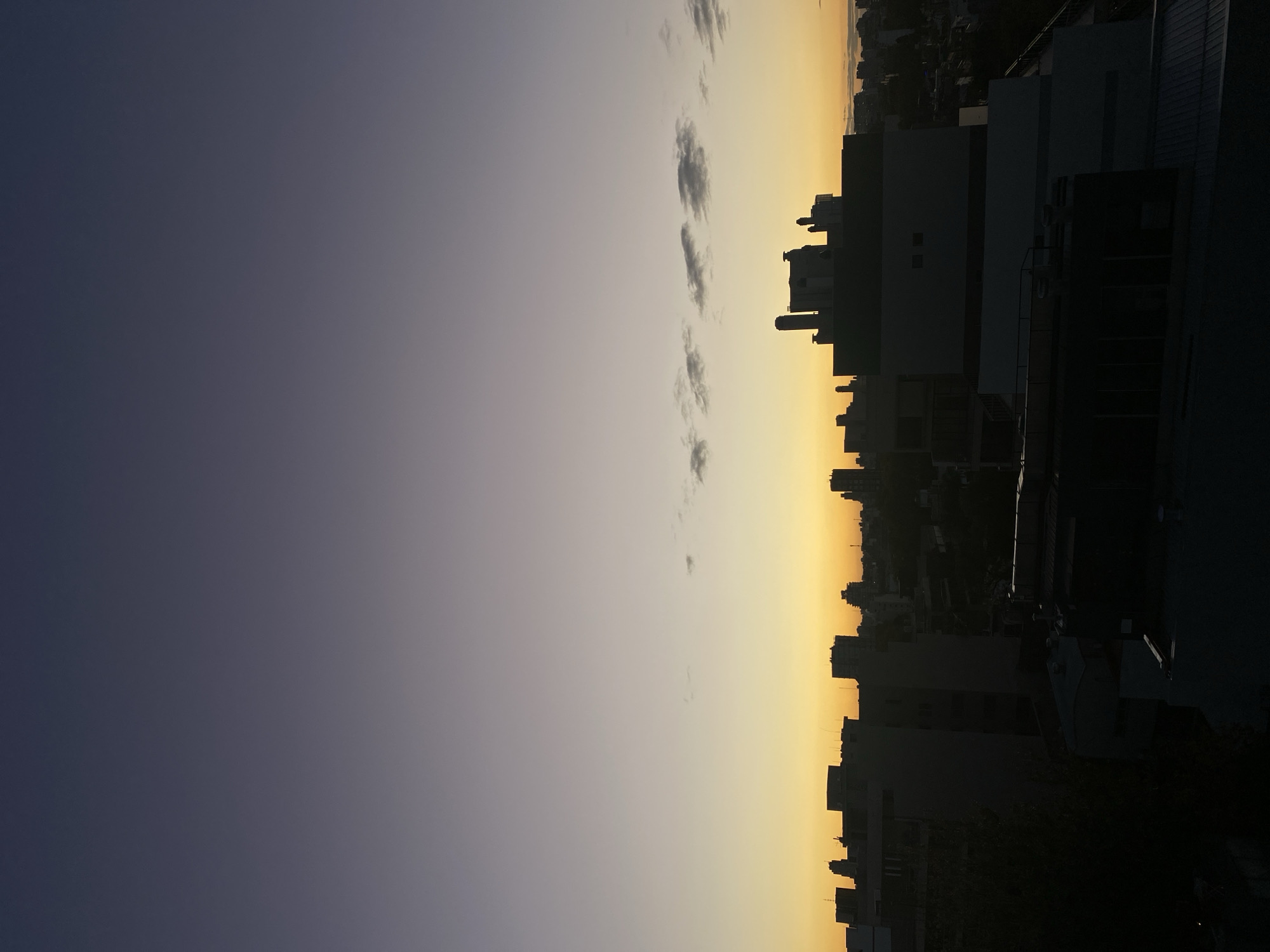 The image shows a cityscape at twilight. The sky transitions from a dark blue-gray on the left to a golden-yellow near the horizon. There are a few scattered clouds in the center of the frame. On the right, buildings of varying heights form a silhouette against the sky. The buildings are mostly dark, with some lighter-colored surfaces visible. The location is likely Buenos Aires, Argentina.