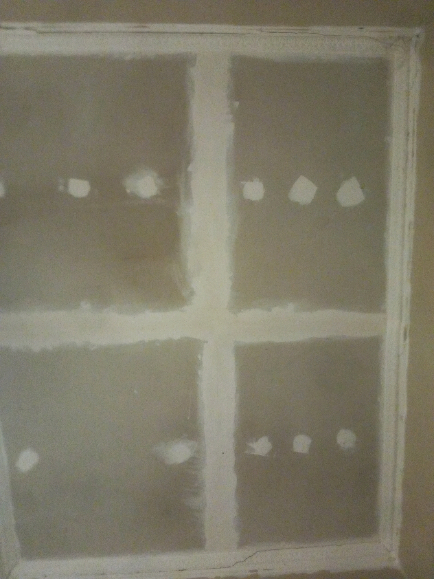 The image shows a close-up, straight-on view of a wall that is undergoing some form of repair or construction. The wall is made of drywall panels that are joined together with tape and mud, creating a grid-like pattern. There are several circular or irregularly shaped patches of drywall mud visible on the surface, suggesting that holes or imperfections are being filled. The overall color of the wall is a neutral, light gray, with the drywall tape and mud appearing lighter and more textured. The wall is framed by what appears to be a white border or trim. The lighting is even, and there are no strong shadows. There are no people or other discernible objects in the image, making it difficult to establish a specific context or location beyond the immediate subject matter of the wall. The image quality is moderate, with some slight blurriness and noise. It is not possible to determine the location in the city of Kasihan, Indonesia, based solely on this image.