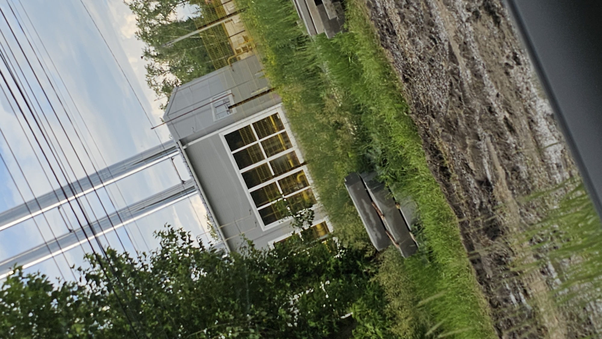 The image depicts an industrial area with a gray building, possibly a small factory or utility structure, with large windows. Two tall, metallic chimneys rise from the roof of the building. The building is surrounded by green grass, some of which is overgrown. In the foreground, there are stacks of concrete blocks, suggesting ongoing construction or renovation. A dirt road with tire tracks and some standing water is also visible in the foreground. Overhead, power lines crisscross the sky. In the background, there is a yellow fence and more trees. The image appears to be taken in an outdoor, semi-rural or industrial setting. The sky is cloudy. There are no people visible in the image. The location is identified as Kamensk-Uralsky, Russia.