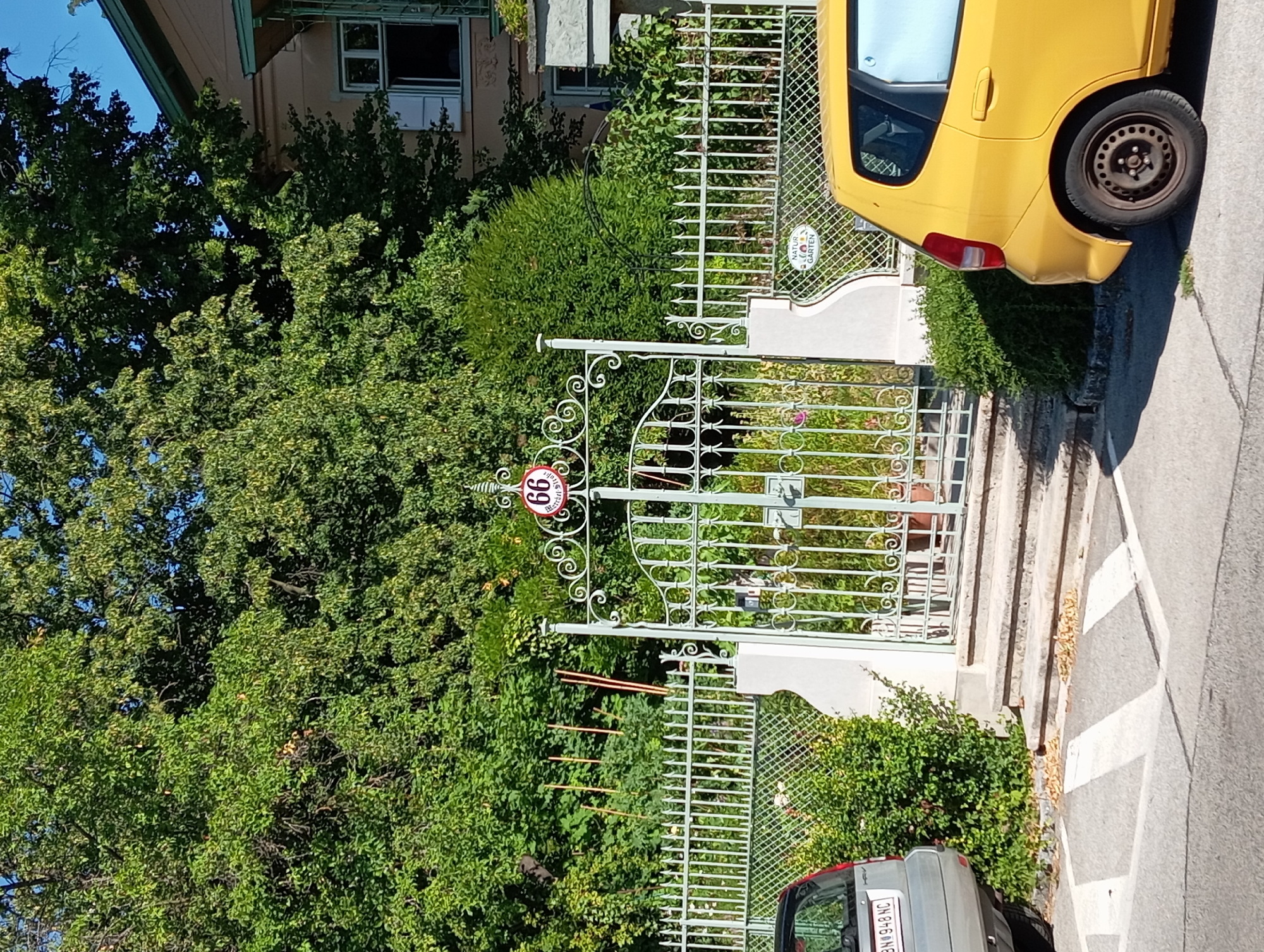 A light green ornate gate stands in the center of the frame, leading to a property with lush green trees and bushes. The gate is adorned with swirling metalwork and a circular sign with the number "66" in red on a white background. To the left of the gate, a silver car is parked, and to the right, a yellow car is partially visible. The cars are parked on a paved area with white markings. The scene is set in Baden, Austria, indicated by the street sign reading "Břeclavská Straße" visible on the gate post, though it is likely a mistranslation or misidentification of a local street name and should be "Břeclavská ulice" if this were in the Czech Republic or Slovakia, or a completely different Austrian street name. The presence of a "NATUR GARTEN" (Nature Garden) sign on the fence further suggests a garden setting. The sky is blue and clear, indicating a sunny day.