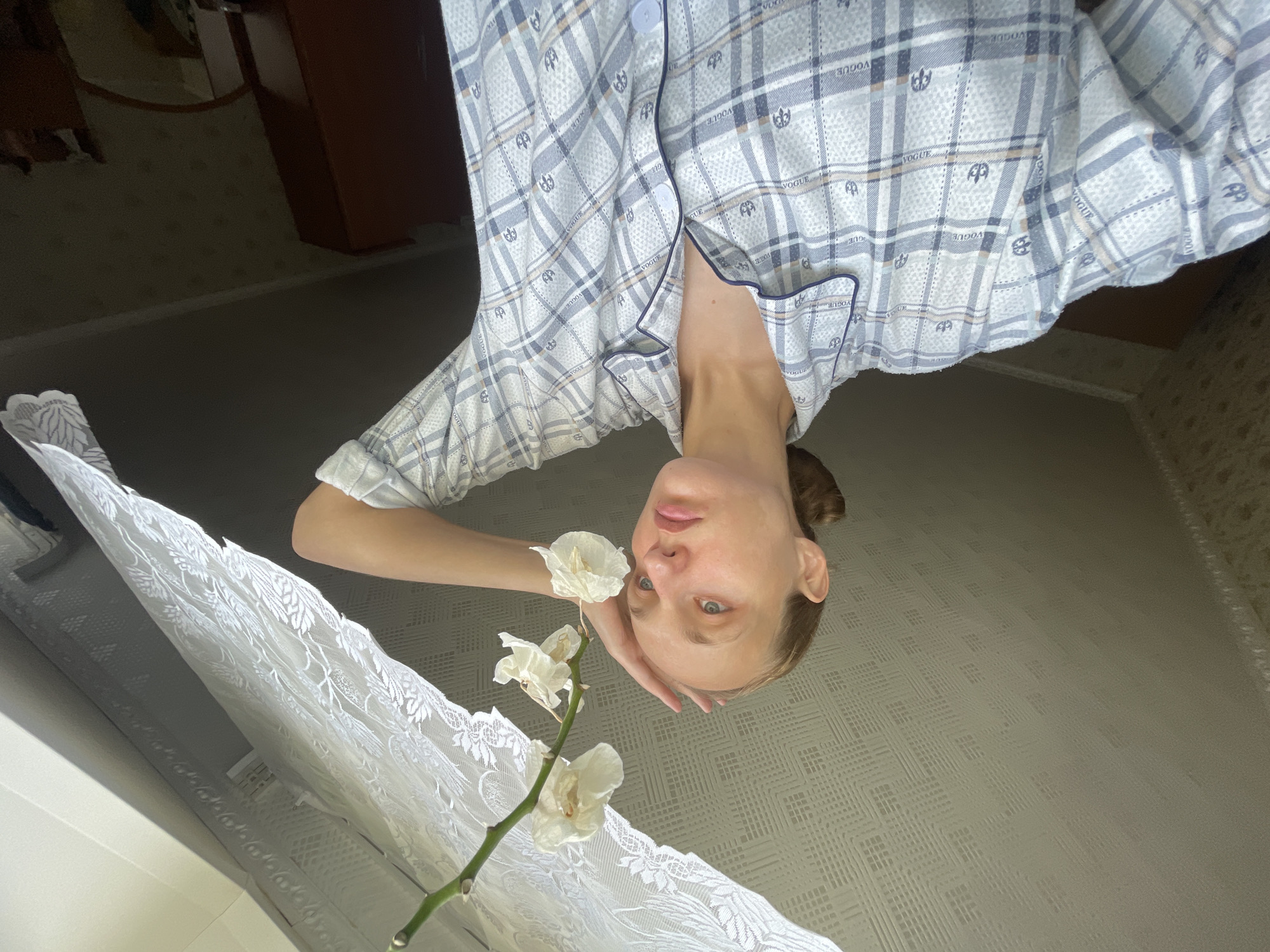 A young woman with blonde hair pulled back in a bun is shown from the chest up, in a light blue and white plaid pajama top. She is looking to her right, with her right hand resting on her forehead. A stem with several wilting white orchids hangs from the upper right corner of the image, extending towards the woman. She is positioned in front of a window covered by a white lace curtain. The ceiling above her has a textured, patterned finish. The background includes a glimpse of a dark brown wooden wardrobe and a mirror, suggesting an indoor residential setting. The overall lighting is soft, possibly from natural daylight filtering through the curtain. No specific landmarks or characteristics of Sharkan, Russia, are identifiable in the image.