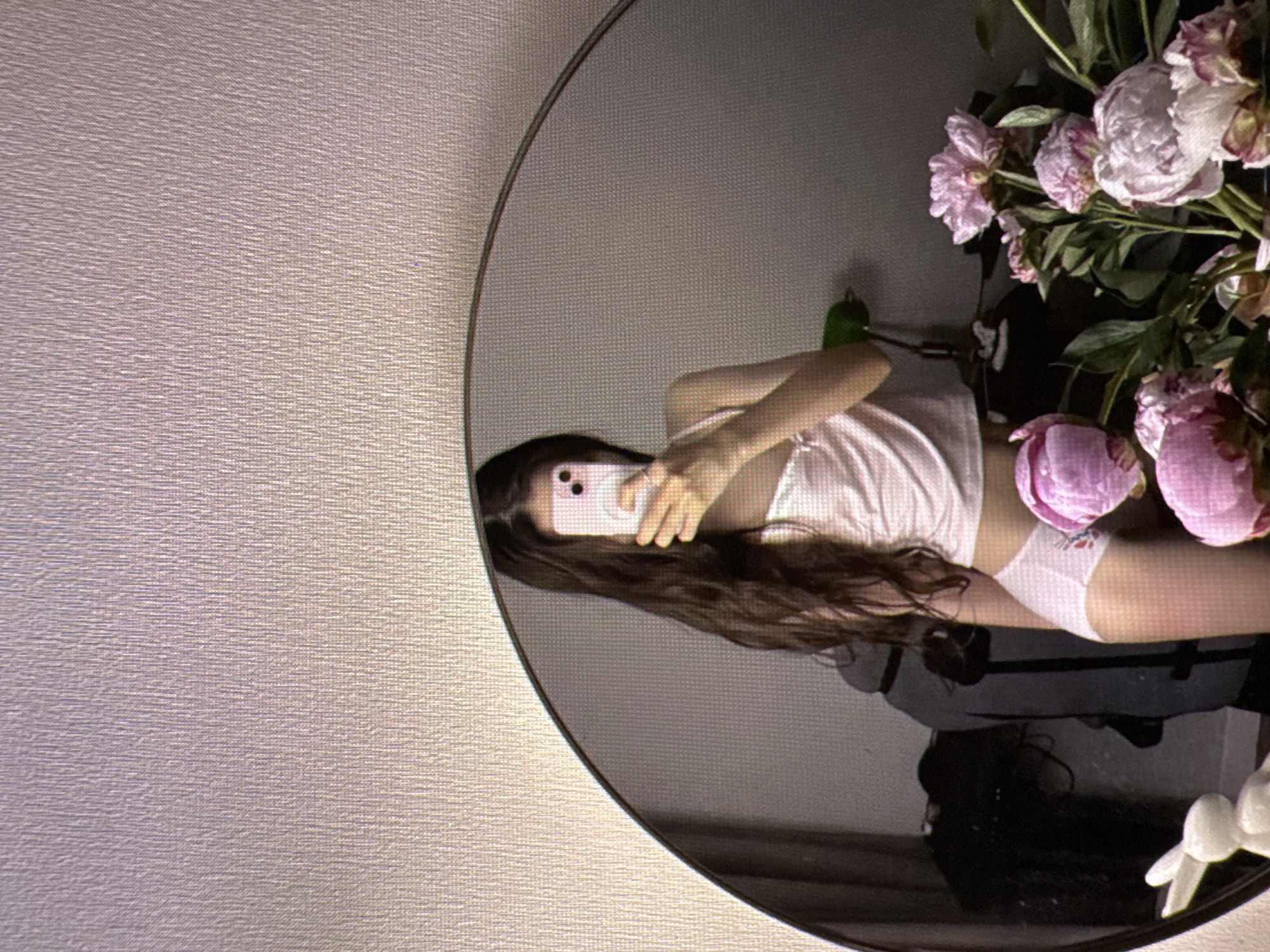 In the city of Castelo Branco, Portugal, a person is captured in a mirror selfie. The scene is indoors, with a textured wall in the background. The person is holding a pink smartphone, taking a picture of themselves reflected in a large, round mirror. They are wearing a light pink tank top with spaghetti straps and white bottoms with a floral pattern. Their long, dark hair falls around their shoulders. In the foreground, to the right of the mirror, a bouquet of pink peonies is arranged. To the left of the mirror, a white ceramic bunny figurine is visible. The overall environment suggests a personal and intimate setting.