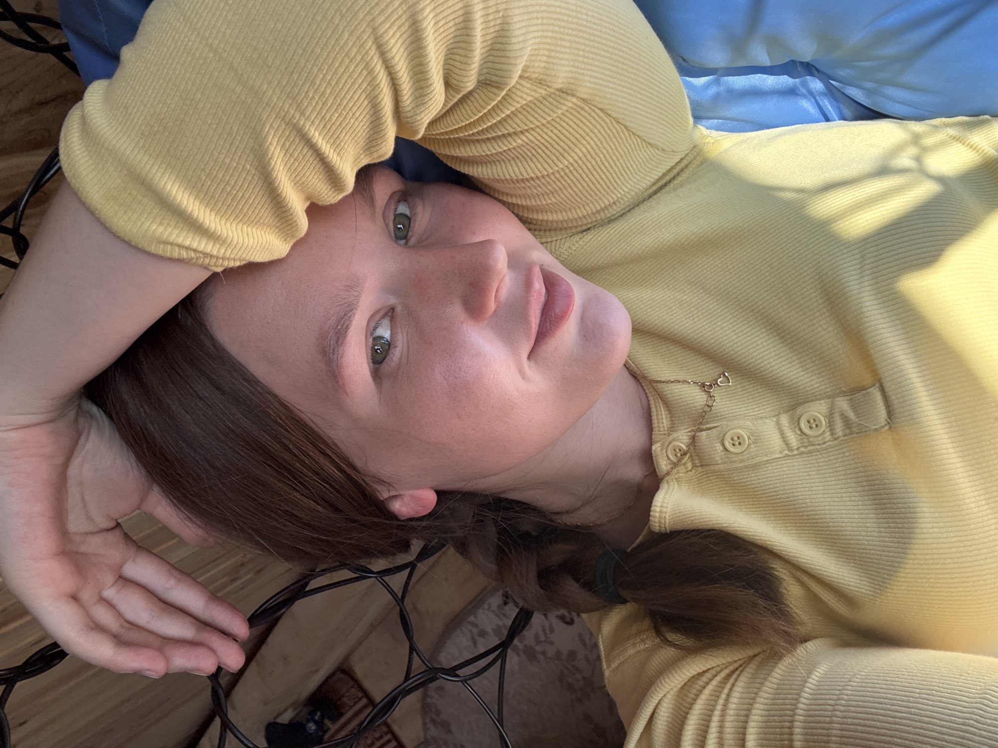 The image depicts a light-skinned female individual, estimated to be in her late teens or early twenties, lying down indoors in Italmas, Russia. She has medium-long, reddish-brown hair, with a braid visible on the left side secured by a dark green tie. Her green eyes are open, and she offers a subtle smile directly towards the camera. She is dressed in a long-sleeved, ribbed yellow Henley shirt with a four-button placket and a gold-colored necklace featuring a small heart-shaped charm. Her right arm is bent and raised, with the hand resting near her head, partially obscured by numerous tangled black electrical wires. The background visible on the left side features light brown wooden paneling and a portion of a patterned brown or beige rug. On the right, a section of blue satin or silky fabric is present, consistent with bedding or upholstery. The scene is illuminated by natural light, casting noticeable shadows across the subject's shirt and the blue fabric.