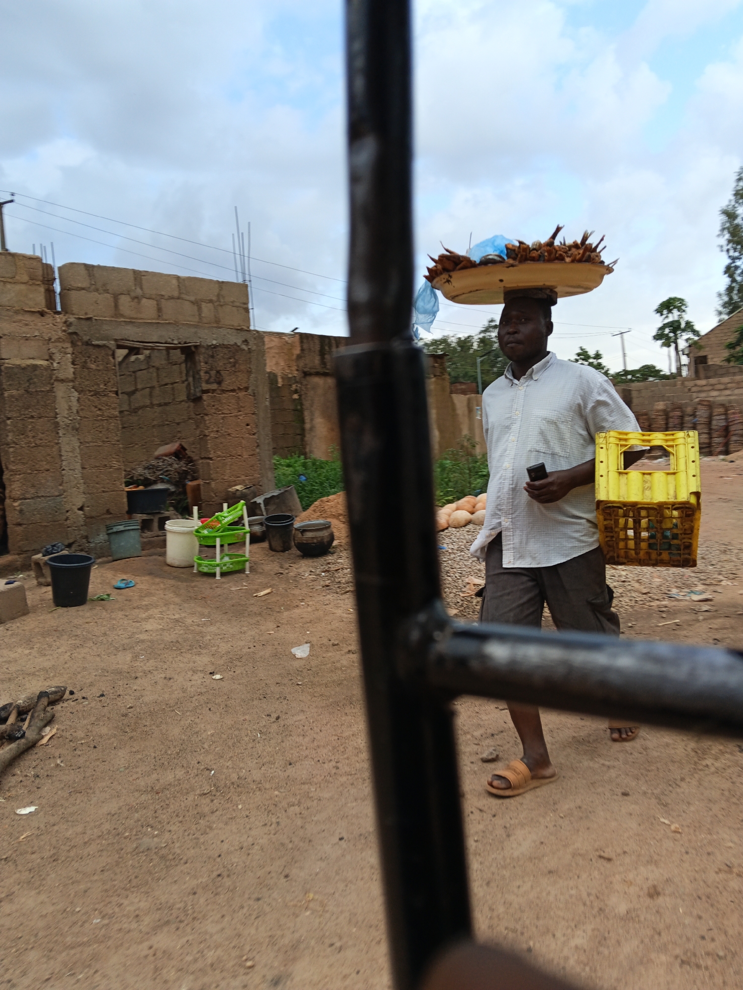 A man walks across a dirt path in an outdoor area, carrying a tray of food on his head and a yellow plastic crate in his hand. He is wearing a light-colored checkered shirt and dark shorts, with tan sandals on his feet. The background shows a partially constructed building made of concrete blocks, some green vegetation, and a cloudy sky. The scene appears to be in a semi-urban or rural setting.