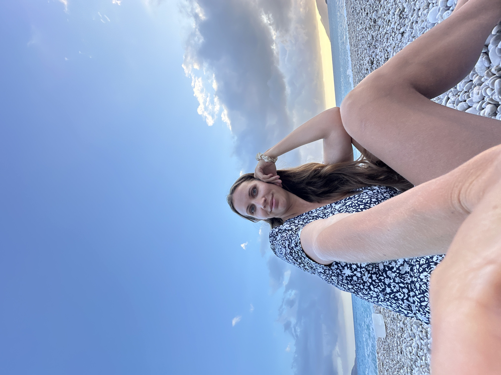 A woman is photographed at an angle, likely a selfie, on a pebble beach in Fethiye, Turkey. The foreground is dominated by the woman's outstretched arm and legs lying on the white pebbles. She is wearing a dark dress with a light floral pattern. Her long, brown hair flows beside her face, and she looks towards the camera. In the background, the sky is a mix of blue and overcast clouds, with a sliver of lighter sky at the horizon. The sea is visible between the horizon and the pebbles.