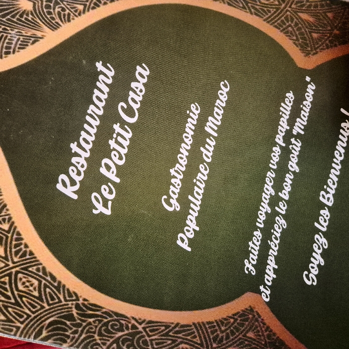 The image features a promotional design for "Restaurant Le Petit Casa," highlighting its Moroccan cuisine with the phrase "Gastronomie Populaire du Maroc." The text invites customers to "Faites voyager vos papilles et appréciez le bon goût 'Maison,'" which translates to "Let your taste buds travel and enjoy the good taste of 'Home'." The bottom line reads, "Soyez les Bienvenus!," meaning "Welcome!". The design is printed on a surface with an intricate, possibly Moroccan-inspired border pattern.