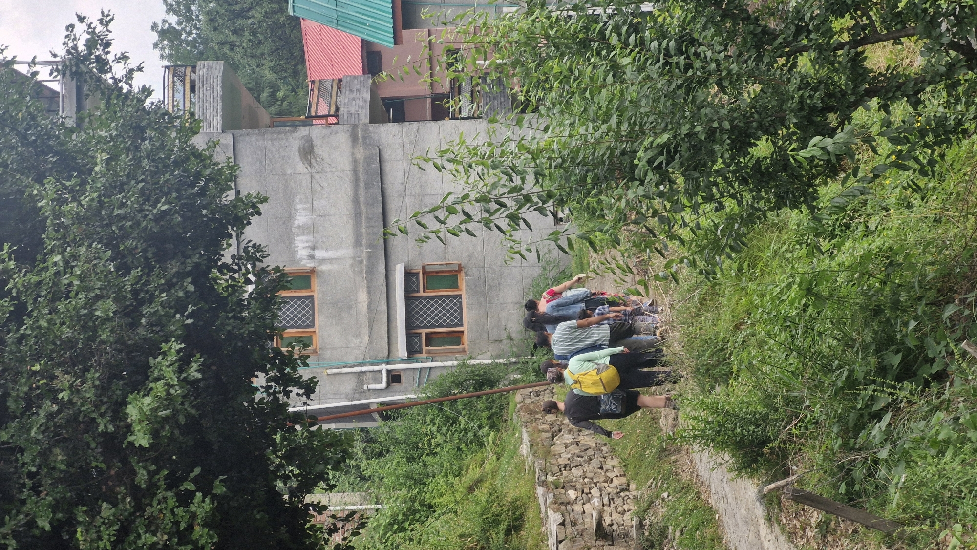 In Vashisht, India, a group of seven people walks along a dirt path beside a stone wall and lush green vegetation. A concrete building with multiple windows is visible behind them, with a balcony on an upper floor. The path leads uphill and curves to the left. The people are dressed in casual clothing, and some are carrying backpacks. The scene appears to be a daytime outdoor setting.