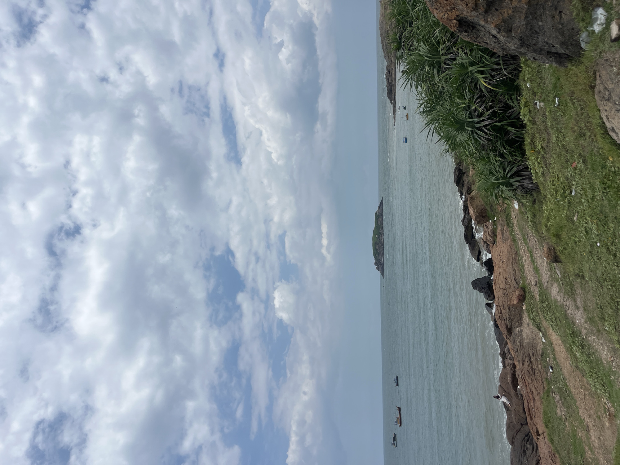 The image captures a coastal scene in Mui Ne, Vietnam. The foreground features a rocky, grassy embankment with scattered litter. Several large, dark rocks are visible, some with a reddish hue. Dense, spiky green vegetation grows on the right side of the frame. A narrow dirt path winds down towards the water.
The middle ground shows a rocky shoreline with waves lapping against the stones. In the water, several small fishing boats are dotted across the sea. One individual, dressed in white, is seated on the rocks near the water's edge.
The background consists of the calm sea and a distant, small, green island. The sky is partly cloudy, with a mix of white cumulus clouds and patches of blue sky. The overall lighting suggests it is daytime, possibly late morning or afternoon.