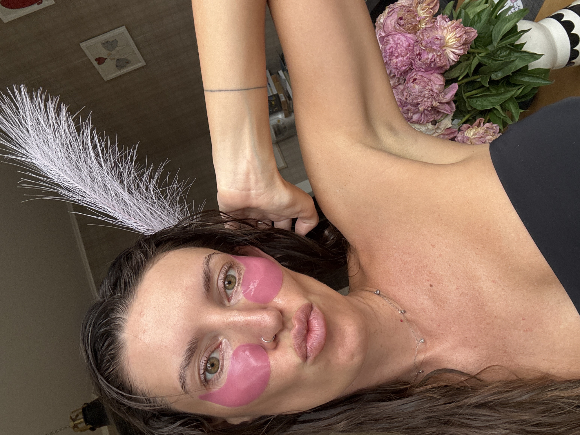 The image shows a medium close-up of a woman with long, dark, wet hair, wearing pink under-eye patches and a black strapless top. She has a silver nose ring and a delicate necklace. Her right arm is raised, with a faint line tattoo visible on her forearm. Behind her, on the right side of the frame, is a vase of wilted pink peonies. The background appears to be an indoor setting with a patterned wallpaper and framed artwork on the wall. A white feather plume is positioned near her head. The lighting suggests it is daytime. The specific location within Moscow, Russia, cannot be determined from the visual information provided.