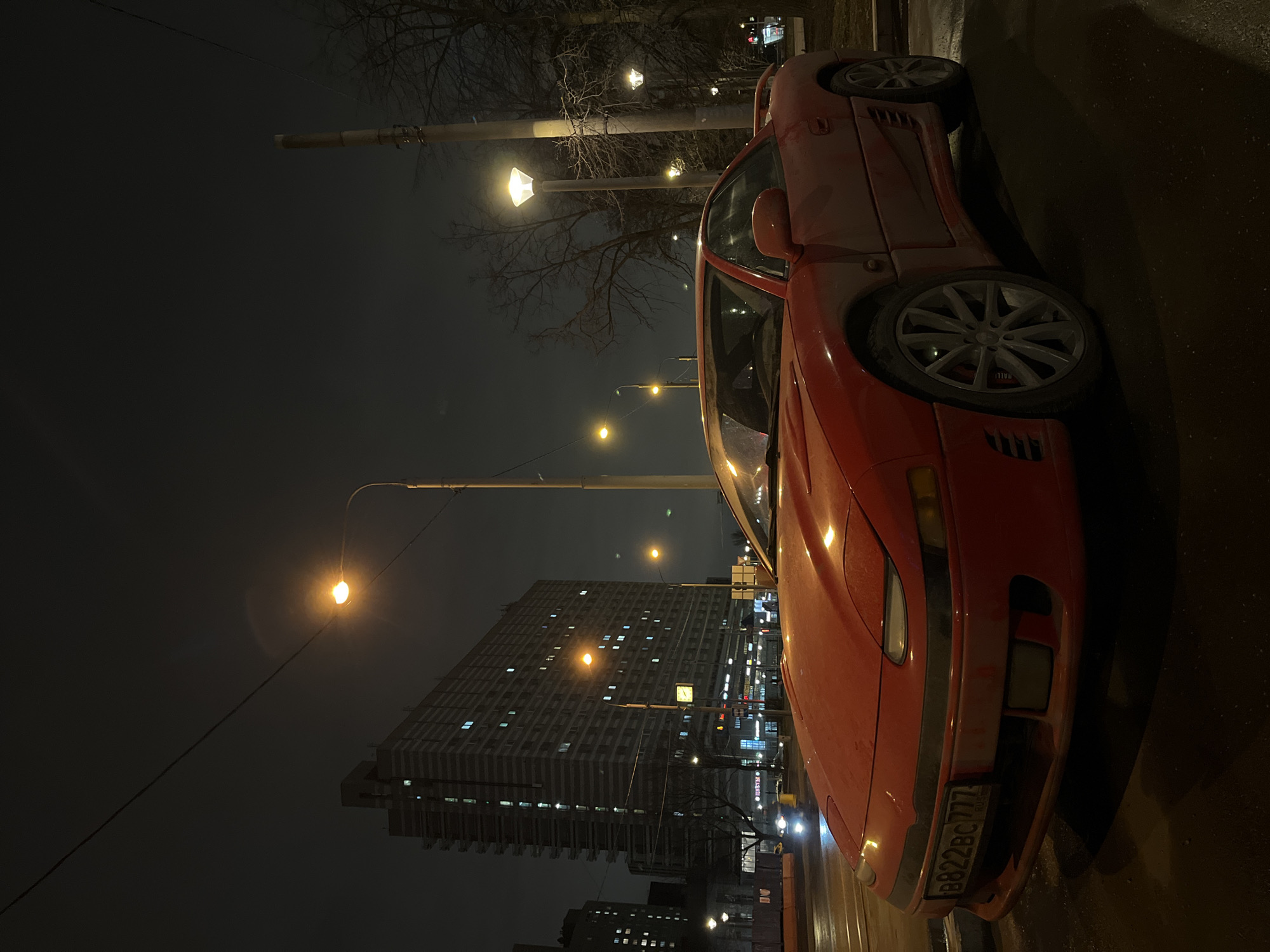 A red Mitsubishi GTO coupe with registration plate B822BC777 is parked on a street in Moscow, Russia at night. A tall, high-rise apartment building with lit windows stands behind streetlights on the opposite side of the street. The pavement is wet.