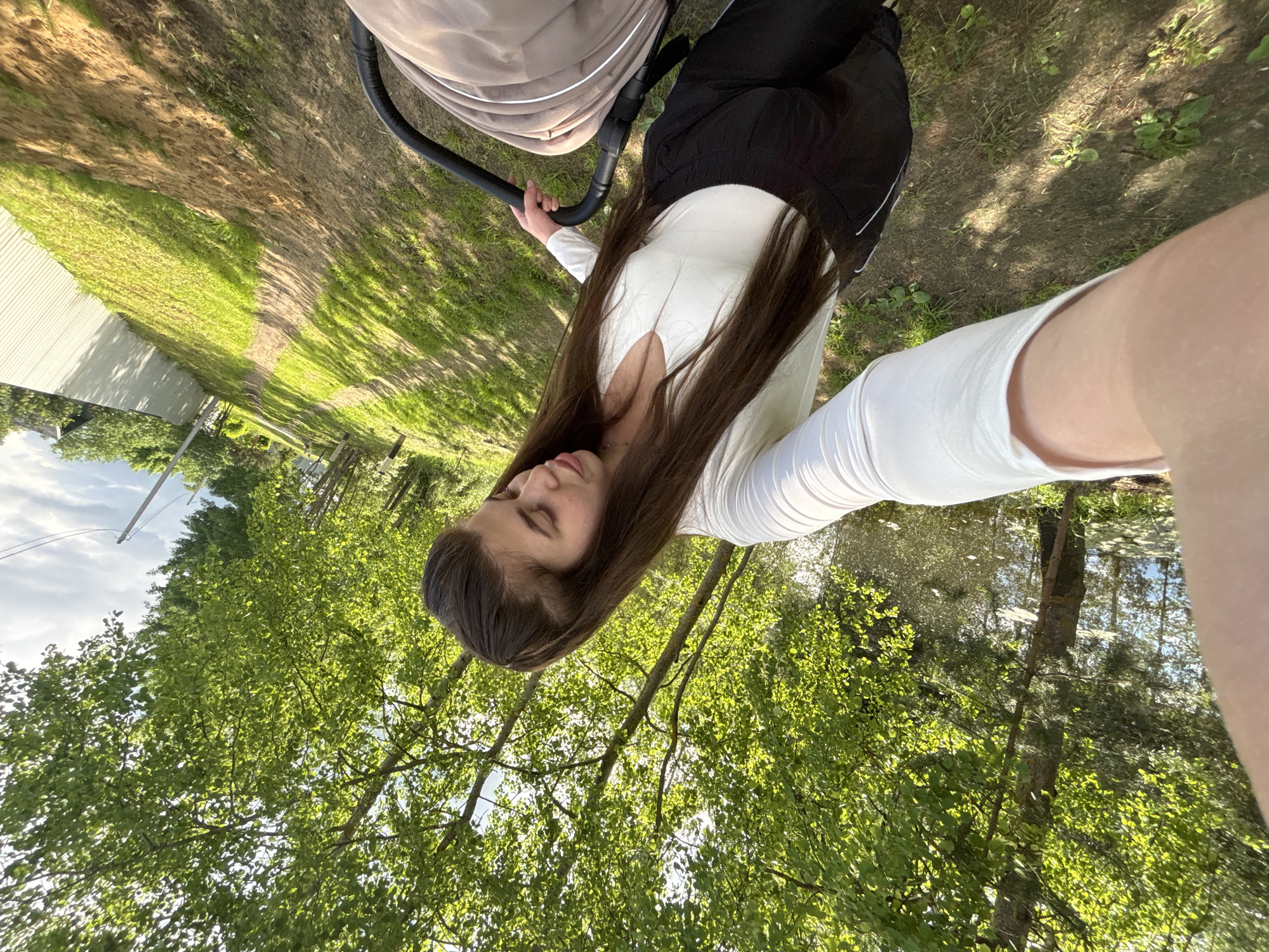 A young woman is outdoors, holding a baby stroller. She has long, dark hair and wears a long-sleeved white shirt and black pants. The stroller is tan-colored. The background shows a grassy area with trees. The location appears to be a park or natural area. Based on the prompt, the location is in the city of Мещёры, Russia.