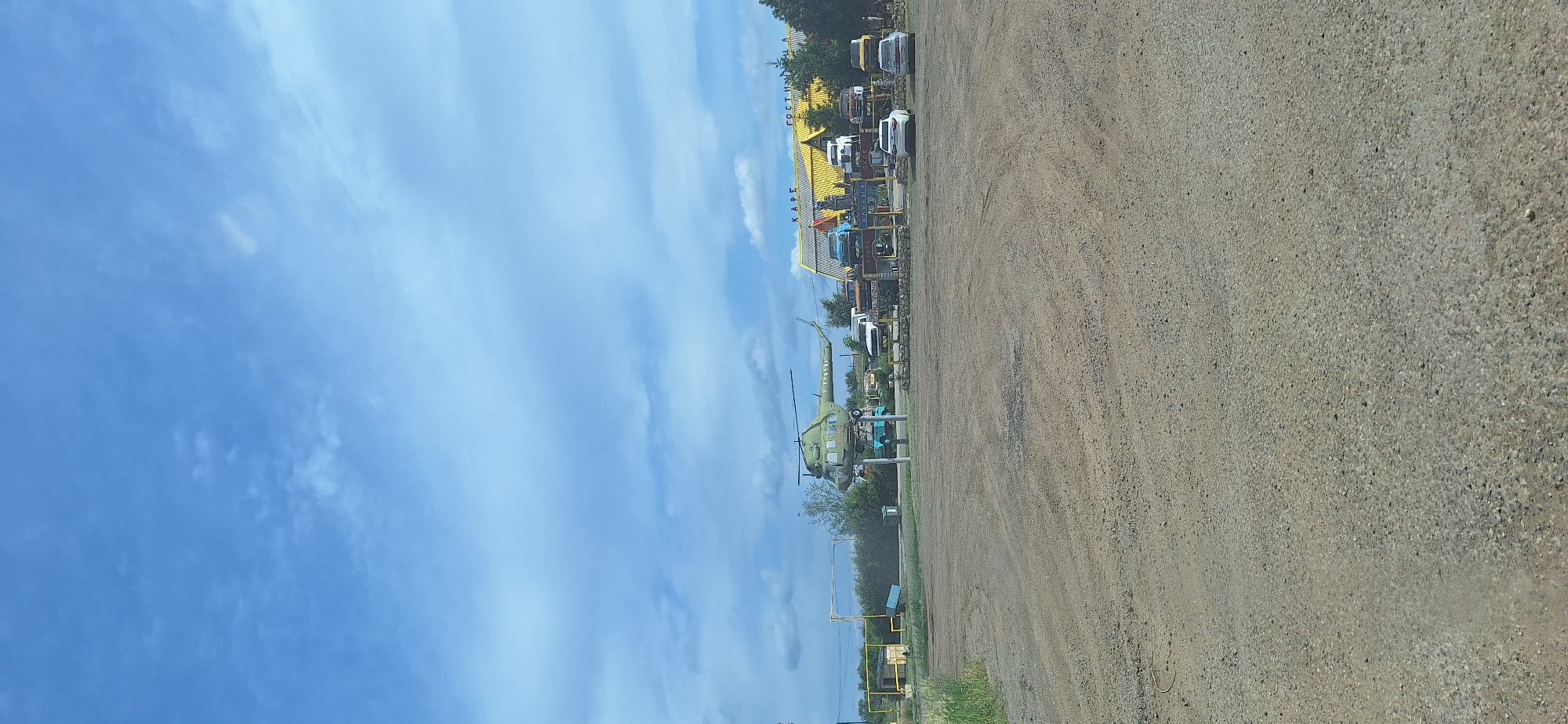 The image shows a desolate landscape under a cloudy sky. In the foreground, a dirt lot stretches towards a collection of buildings and vehicles in the midground. A green helicopter is prominently displayed, mounted on a platform. To its right, a long, yellow building with "КАФЕ" and "ГОСТИНИЦА" written on it suggests it's a cafe and hotel. Several cars are parked in front of and around the buildings, some on elevated platforms. The overall scene appears to be a roadside establishment in a rural or undeveloped area.