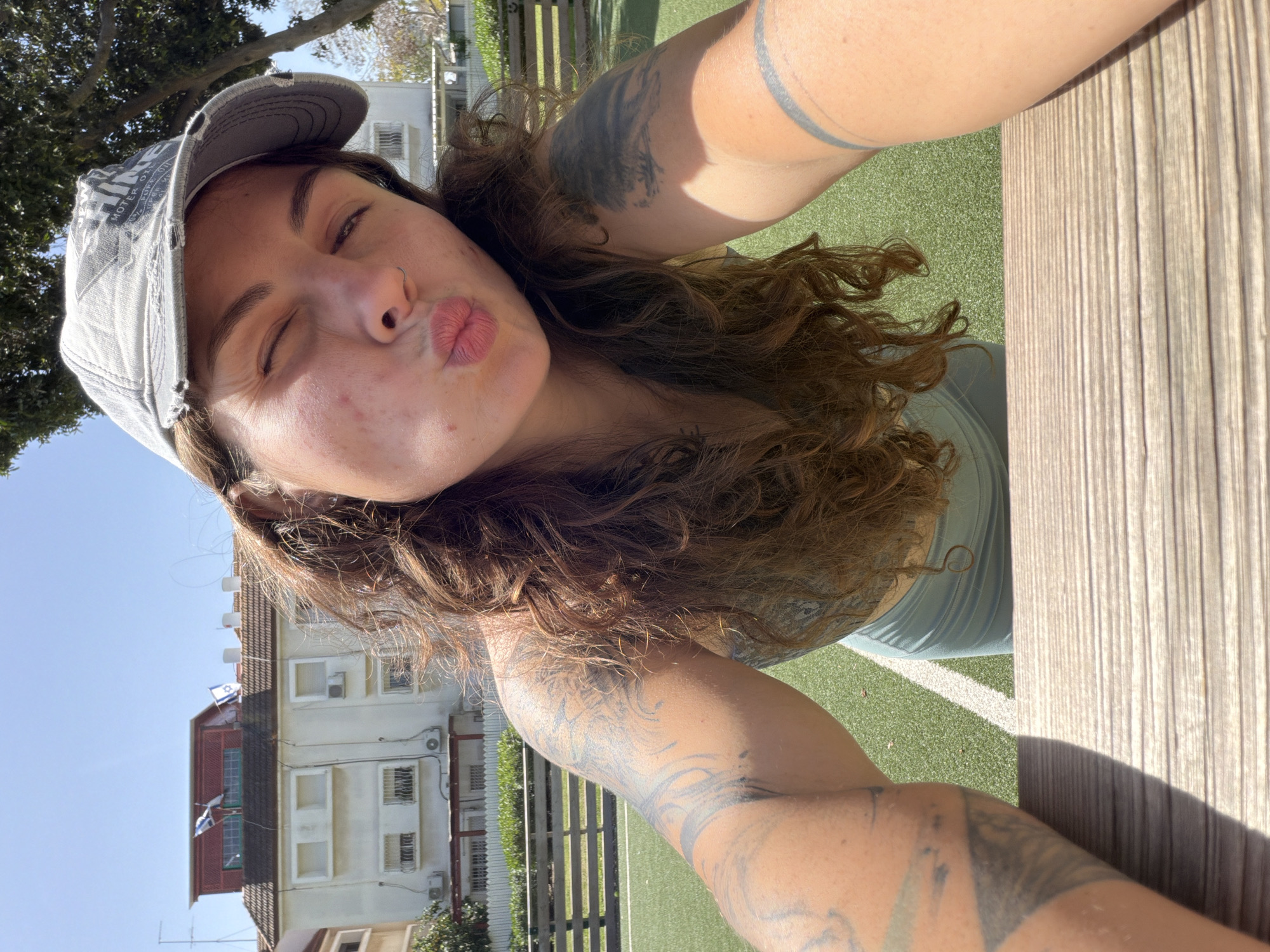 A woman is taking a selfie outdoors, near a wooden table. She is wearing a gray baseball cap and a light blue top. Her left arm and right forearm are visible and covered in tattoos. Her hair is curly and brown. She is puckering her lips and closing her eyes in a kiss gesture. In the background, there's a building with white walls, a red roof, and several windows. The flag of Israel is visible on the building. The location is Kiryat Bialik, Israel, based on the Israeli flag. There is a green lawn and a fence in the background. The sky is bright and clear.