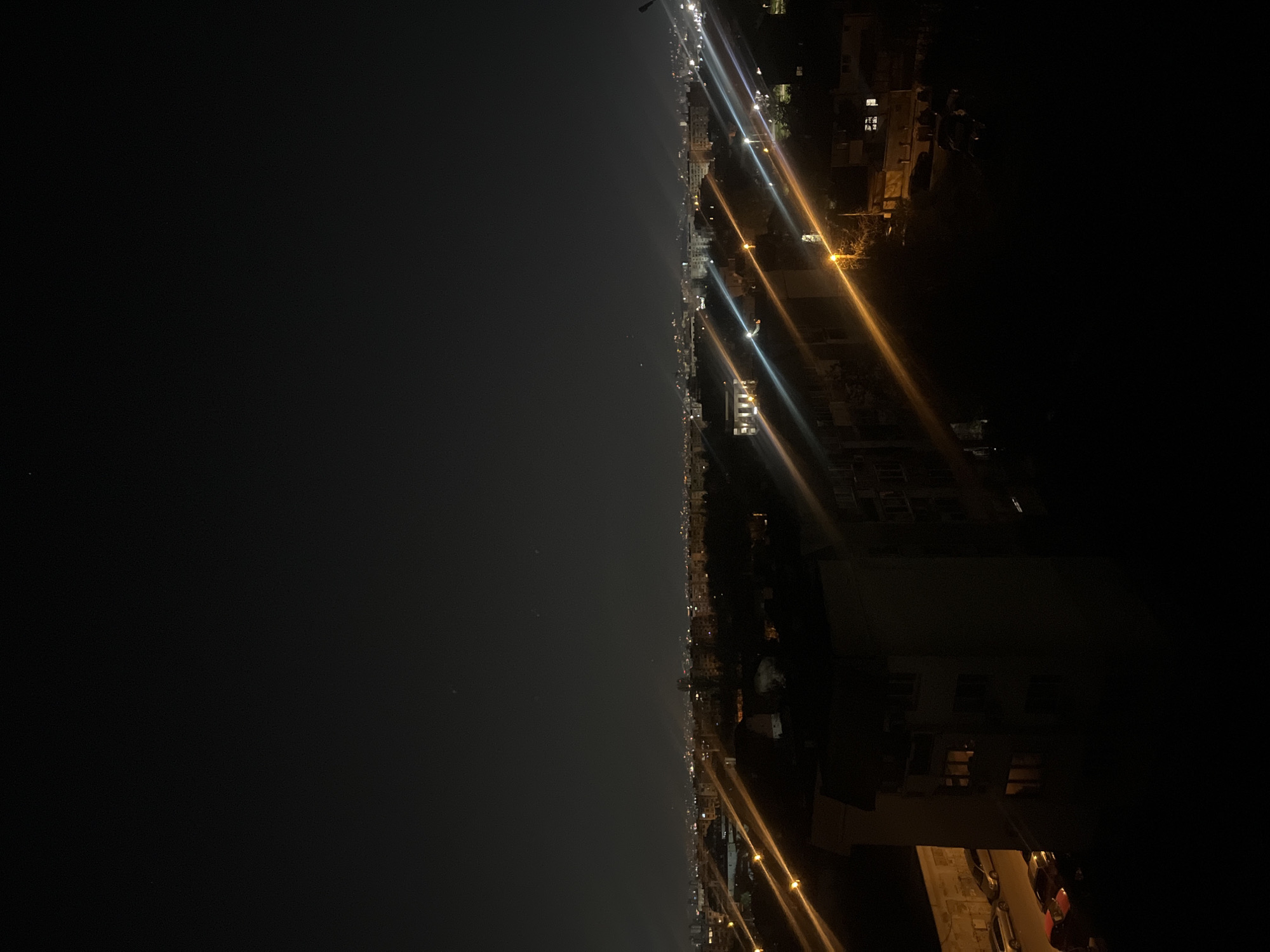 This media file presents a panoramic view of a city at night, likely Sofia, Bulgaria, stretching across the horizon. The image, when viewed in its intended horizontal orientation (rotated 90 degrees counter-clockwise from the provided upload), depicts a vast expanse of dark, clear night sky in the upper portion, dotted with a few faint specks of light that could be stars or distant urban illumination.
Below the dark sky, a long, continuous line of urban development is brightly lit, extending into the distance. This cityscape is characterized by numerous artificial lights, predominantly yellow-orange from streetlights and some white or blue from other sources like building interiors or vehicle headlights. These lights create prominent, elongated streaks across the frame, suggesting either a long exposure photograph capturing moving vehicle traffic or significant lens flare from the intense light sources.
Dimly discernible multi-story buildings line the illuminated strip, with several showing windows glowing with internal lights. These structures indicate a dense urban environment. The lower-right corner of the horizontally oriented image shows a closer view of a building or structure, providing some foreground detail.
The scene conveys the vibrant activity of a city after dark under clear weather conditions, without specific people or discernible text being visible. The long light trails strongly imply the presence of active roads and continuous traffic flow.