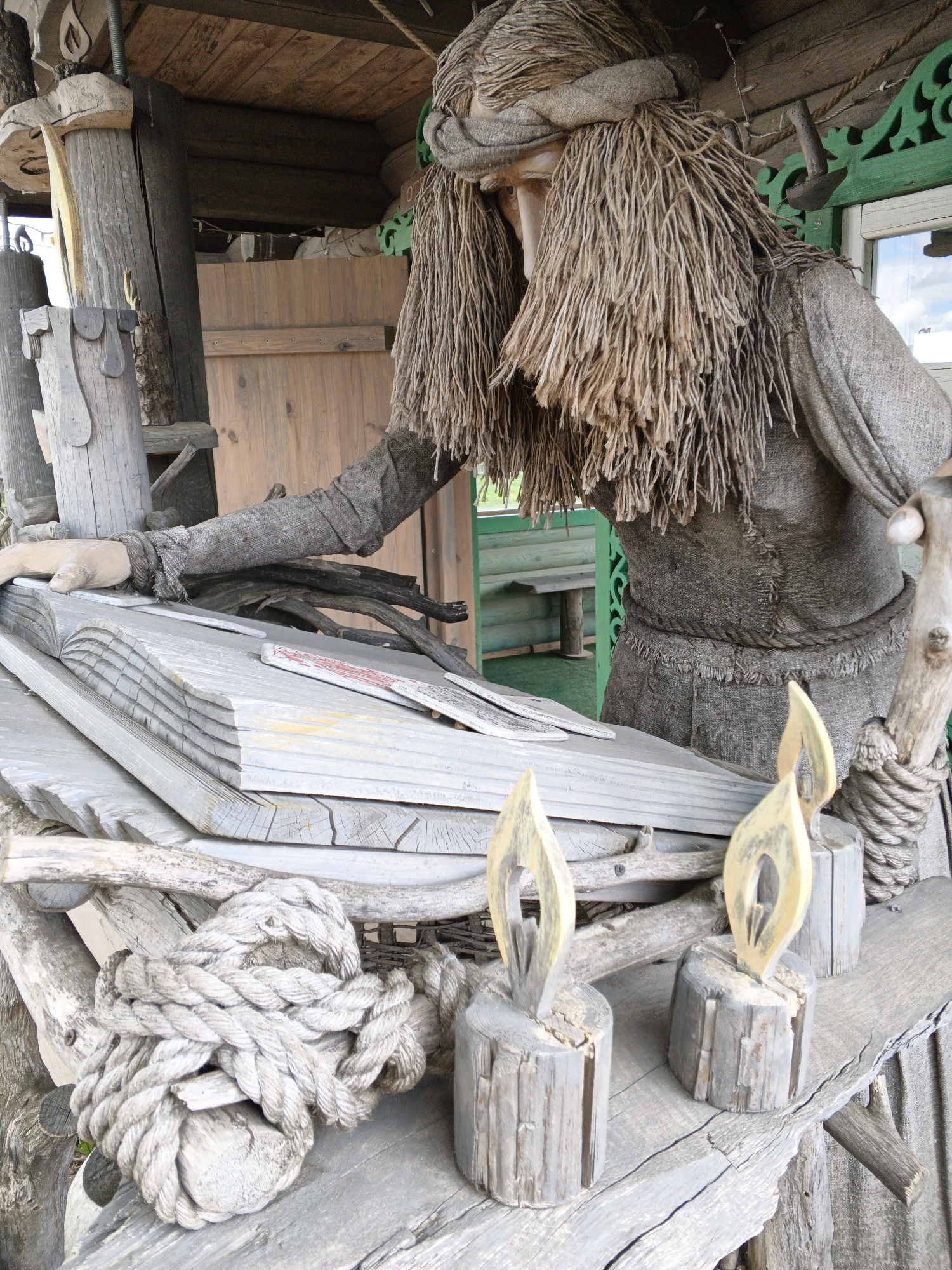A life-sized mannequin, resembling an old woman or a mystical figure, is the central subject of this image. The figure is clad in a rough, homespun garment that appears to be made of burlap or a similar natural fiber, and its head is covered with a bandanna-like wrap of the same material. Long, shaggy hair made of straw-like material frames its face.
The mannequin is positioned leaning over a large, open book, intricately carved from weathered wood to simulate pages. Its left hand rests upon the wooden 'pages,' suggesting an act of reading or contemplation. On the wooden surface in front of the mannequin are two carved wooden candles, designed with flame-shaped tops. A thick, coiled rope rests nearby, adding to the rustic aesthetic. Various other pieces of aged wood, including branches and logs, are incorporated into the display.
The setting appears to be a rustic outdoor or semi-outdoor space, possibly a porch or a small wooden structure like a gazebo. In the background, a wooden building or wall is visible, featuring a window with ornate, green-painted gingerbread trim. Through the window, a glimpse of a wooden bench and a bright sky can be seen, indicating it is daytime with clear weather.
The scene creates a tableau evoking folklore or ancient wisdom, possibly depicting a traditional storyteller or a wise woman. No discernible legible text is visible on the wooden book or elsewhere in the image, although some faint, abstract markings are present on the 'pages' and possibly elsewhere as decoration. The overall impression is one of traditional craftsmanship and cultural storytelling, consistent with a setting in rural Belarus.