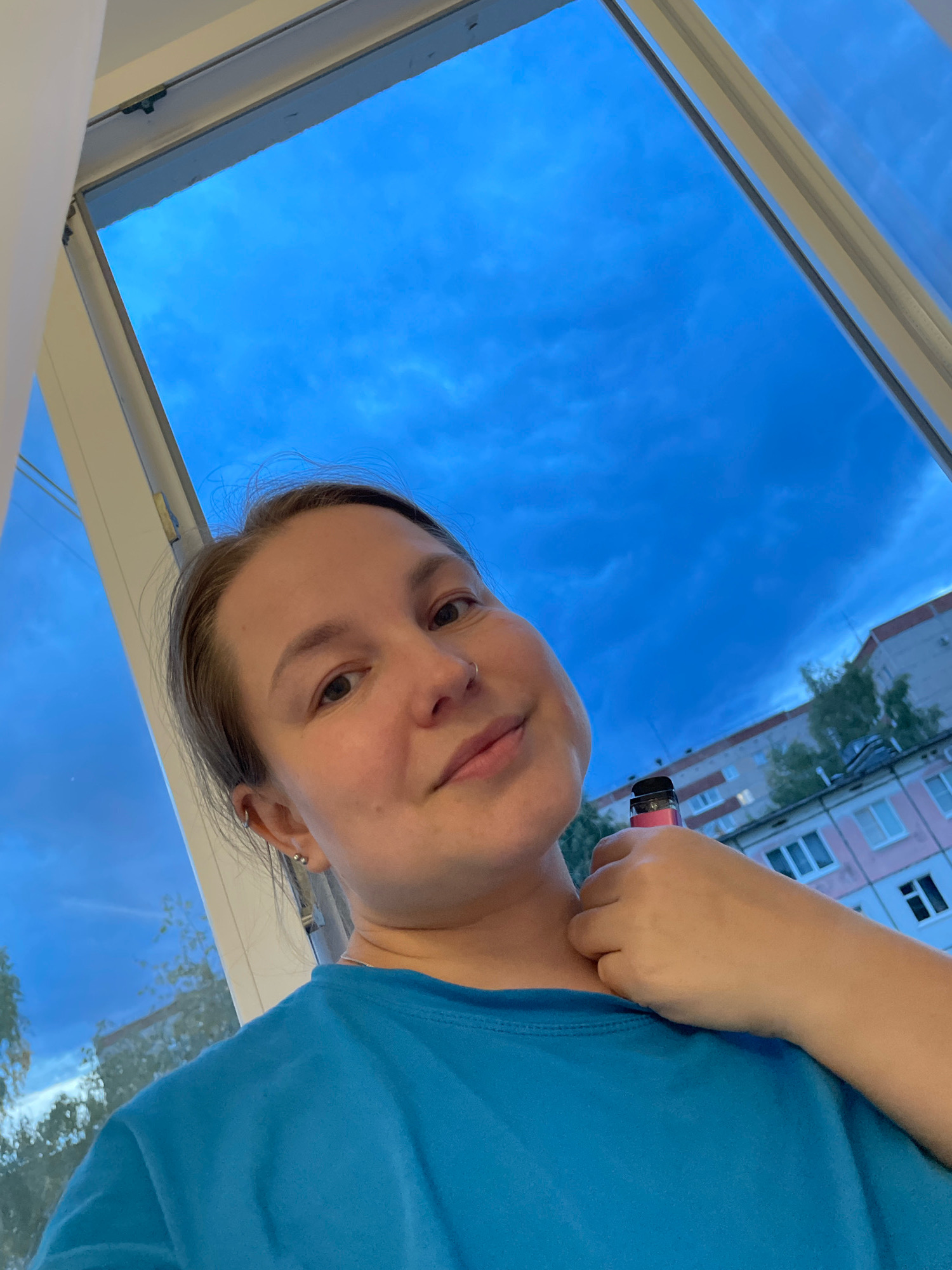 In Izhevsk, Russia, a young woman with light brown hair pulled back is taking a selfie by a large window. She is wearing a bright blue t-shirt and has a nose ring and earrings. Her left arm is resting on her neck, and she is holding a pink vaping device. Outside the window, the sky is a deep blue with scattered clouds, and apartment buildings are visible in the background, some with pink and light gray facades. Trees are also present between the buildings. The lighting suggests it is either early morning or late evening.
