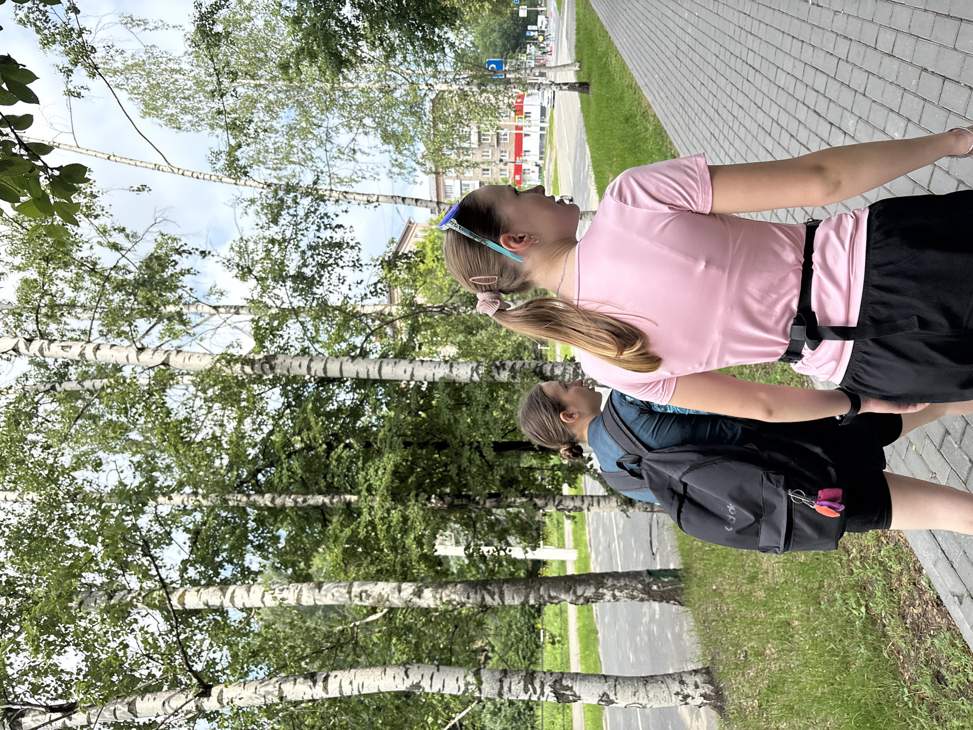 Two young women are walking along a paved sidewalk lined with birch trees on a cloudy day. The woman in the foreground, wearing a pink t-shirt and black shorts, has her back to the camera and is looking to the right. She has her hair in a ponytail with a pink scrunchie and is wearing blue-green sunglasses on her head. The woman behind her is wearing a dark teal t-shirt and black shorts and has a black backpack on. She is looking to her left. Behind the trees, there are buildings and a street with cars. The scene appears to be in a residential area of Izhevsk, Russia, possibly along a street like Udmurtskaya Street or a nearby park, given the prevalence of birch trees and typical Soviet-era apartment buildings visible in the background.
