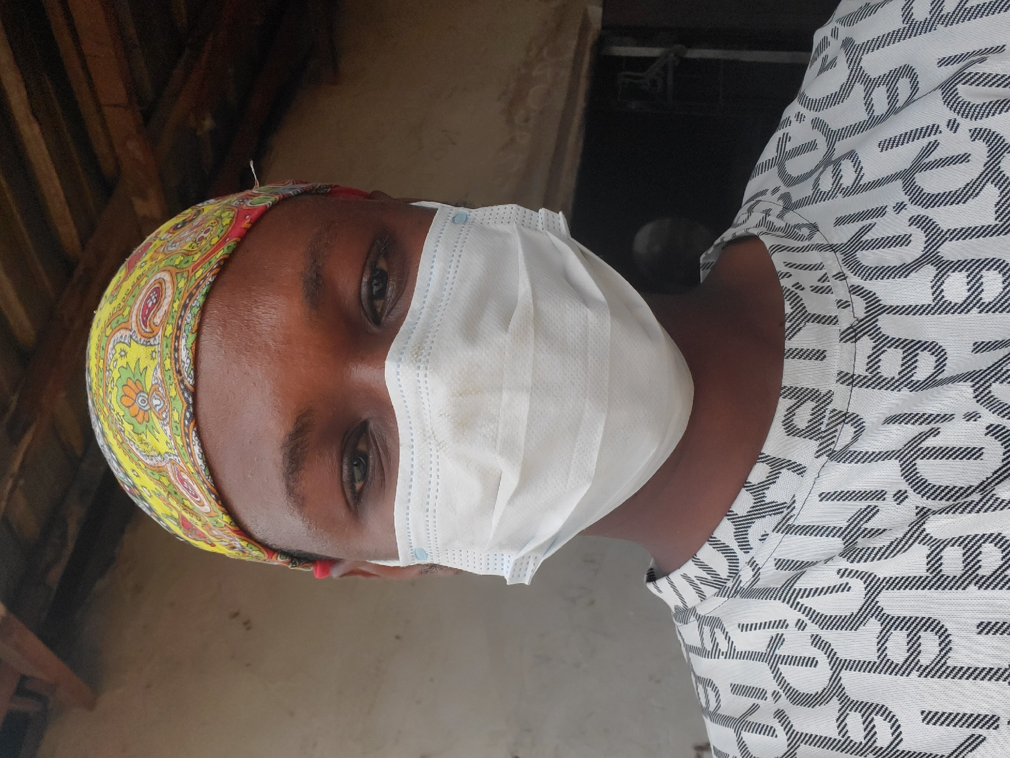 A person wearing a paisley headscarf and a white surgical mask is indoors. The person is wearing a patterned white and black shirt. The location is Mararaba, Nigeria.