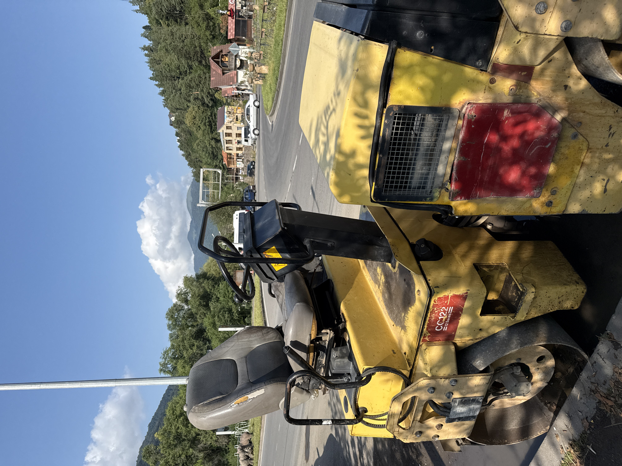 An outdoor, eye-level, vertical shot captures a yellow road roller parked on the side of a road. The roller's worn, gray seat is prominent in the foreground, with a black steering wheel and control levers positioned behind it. The machine's yellow body is visible, with a red sticker on the side that reads "CC122 12 SERIES". Sunlight casts dappled shadows on the roller's surface. In the background, a paved road curves to the right, bordered by a grassy verge and trees. Across the road, a cluster of buildings with red-tiled roofs and colorful facades suggests a town setting. Mountains covered in dense green forest rise in the distance, under a clear blue sky with scattered white clouds. The scene is likely located in Dilijan, Armenia, known for its mountainous terrain and scenic views.