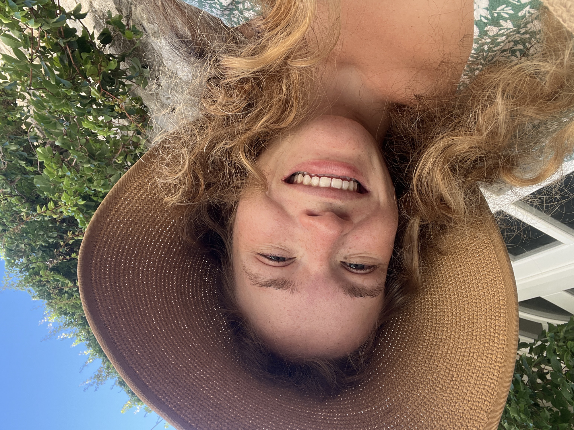 The image is an upside-down, close-up selfie of a young woman taken outdoors in Budva, Montenegro, on a bright, sunny day. The subject is a woman with curly, light brown or blonde hair and freckled skin. She is wearing a wide-brimmed, woven straw hat and a green and white floral-patterned top or dress. Her face, seen from a low angle and inverted, shows a broad, cheerful smile with visible teeth, and her light-colored eyes are slightly squinted in a joyful expression.
The background, visible around her head and hat, consists of lush green foliage from trees or bushes, patches of clear blue sky, and glimpses of a white building structure, possibly a house. The bright lighting suggests it is daytime with good weather. The activity is clearly a selfie, capturing a happy moment. There is no visible text in the image.