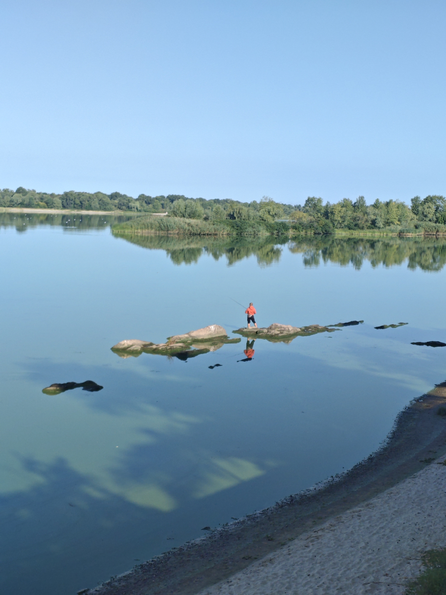 An aerial, eye-level, long shot captures a serene lake scene under a clear blue sky, likely located in Kremenchuk, Ukraine. The foreground features a sandy embankment sloping down to the water's edge, with sparse vegetation and debris. The lake's surface is calm and reflective, mirroring the lush green treeline and scattered clouds on the horizon. In the middle ground, a person in an orange jacket stands on a cluster of rocks, fishing with a rod. Their reflection is visible in the water, as are the dark shadows of submerged rocks or objects. Several other dark, irregular shapes also break the water's surface further out. The far bank is lined with dense deciduous trees, creating a natural, tranquil backdrop. The overall atmosphere is one of peaceful solitude and connection with nature.