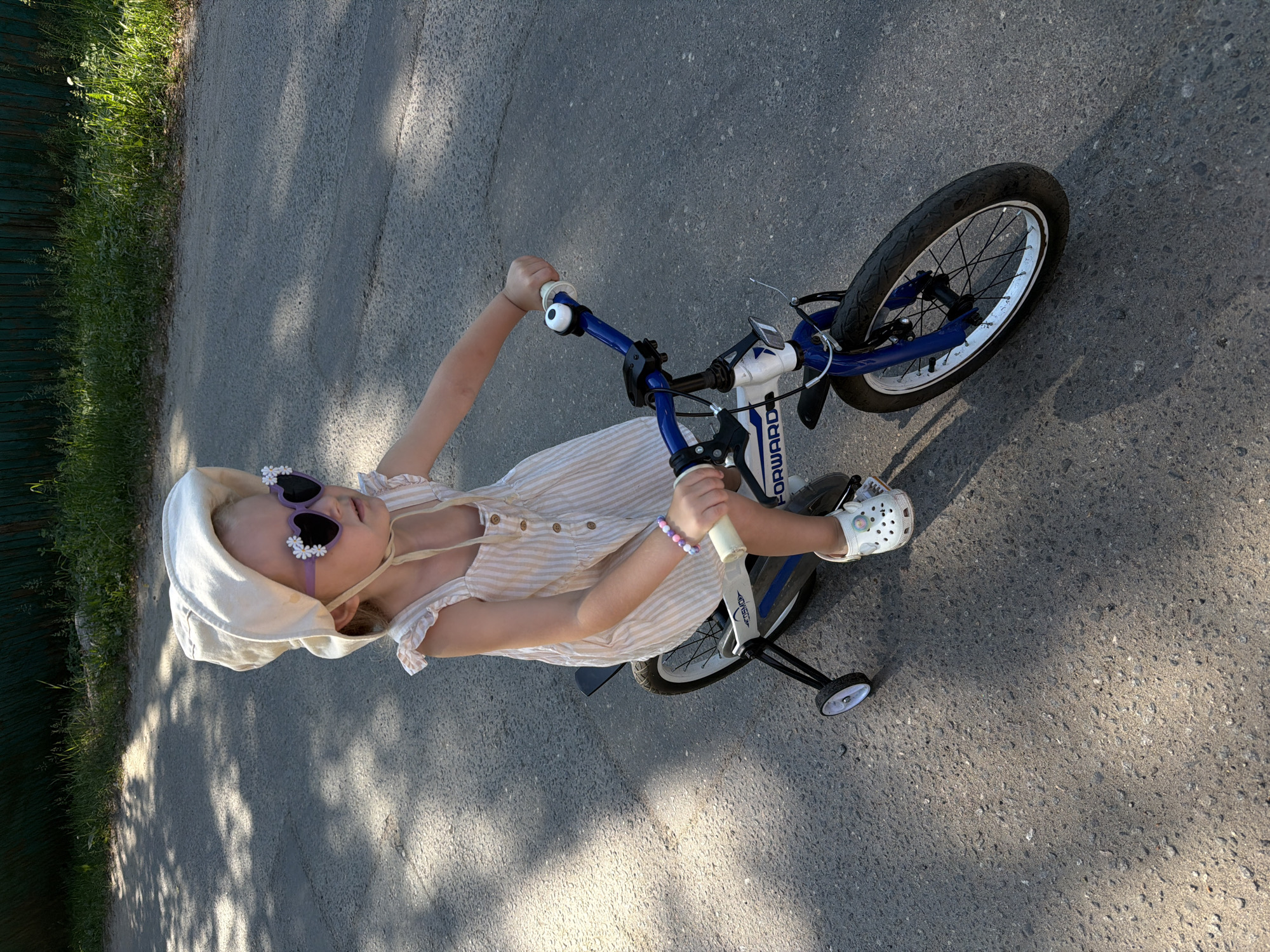 A young girl wearing a sun hat and heart-shaped sunglasses rides a bicycle equipped with training wheels on a paved road. The girl is dressed in a light-colored, striped dress and white clogs. She is holding onto the handlebars, and her left hand is adorned with a beaded bracelet. The bicycle is blue and white, with the brand "FORWARD" visible on the frame. The background features a green fence and grass, suggesting an outdoor setting, likely in Udelnaya, Russia, given the context. The lighting indicates it is daytime, with shadows cast on the road by overhead foliage.