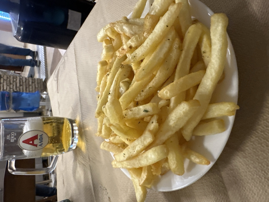 The image, oriented on its side, captures a close-up of a casual dining experience, likely in Nea Erithrea, Greece. The main subjects are a white plate piled high with golden-brown french fries, and a clear glass mug containing light-colored beer with a frothy head.
The fries appear freshly cooked and are lightly seasoned with visible green specks, possibly dried herbs like oregano or parsley. The beer glass prominently features a red 'A' logo and the Greek word "ΑΛΦΑ" (Alfa), identifying it as a popular Greek beer brand. Below the logo, "0.5 L" is visible, indicating the volume of the beer.
The food and drink are placed on a table covered with a light, possibly beige, tablecloth or large napkin. The background is slightly blurred but offers glimpses of the setting. To the left, the lower halves of people's legs and feet are visible, suggesting other individuals in the vicinity. Below them, blue objects or containers are partially seen. A dark, rectangular piece of equipment, possibly a speaker or light fixture, is suspended above the background figures. What appears to be brickwork or stone is also visible in the background, hinting at an outdoor or rustic architectural style for the establishment. The lighting appears bright and warm, suggesting daytime or early evening. The scene portrays a relaxed atmosphere, typical of a casual restaurant or tavern.