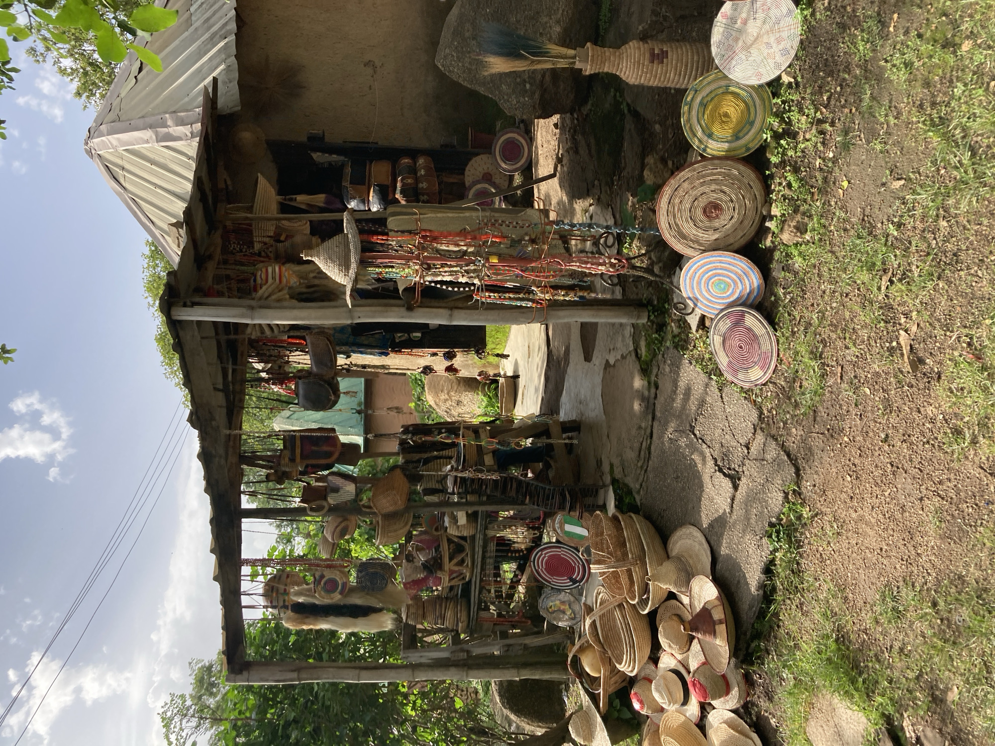 This media file showcases a market stall displaying a variety of handcrafted woven items, predominantly baskets and hats, situated in Jos, Nigeria. The stall appears to be an open-air structure with a corrugated metal roof, supported by wooden posts. It is set against a backdrop of what seems to be a rustic, possibly mud-brick building, with large boulders nearby.
The primary subjects are the numerous woven objects. These include a diverse collection of baskets of various sizes and patterns, some with intricate circular designs, others appearing more utilitarian. Several hats, mostly straw, are also visible, some stacked and others laid out. Hanging from the stall are strands of colorful beads and what appear to be other decorative items, possibly jewelry or adornments. A tall, woven vase holding a broom or fan-like object is prominently placed on a rock.
The scene suggests a market or a vendor selling artisanal goods. The arrangement of the items indicates that they are for sale. The time of day appears to be daytime, with bright sunlight casting shadows, suggesting it's likely morning or afternoon. The sky is partly cloudy, with patches of blue. The ground is a mix of dirt, gravel, and patchy green grass. Lush green foliage from surrounding trees is visible, indicating a natural, possibly rural or semi-urban setting.
Notable details include the texture and craftsmanship of the woven items, highlighting traditional artistry. The overall impression is one of vibrant local commerce and cultural heritage. There is no visible text in the media file.