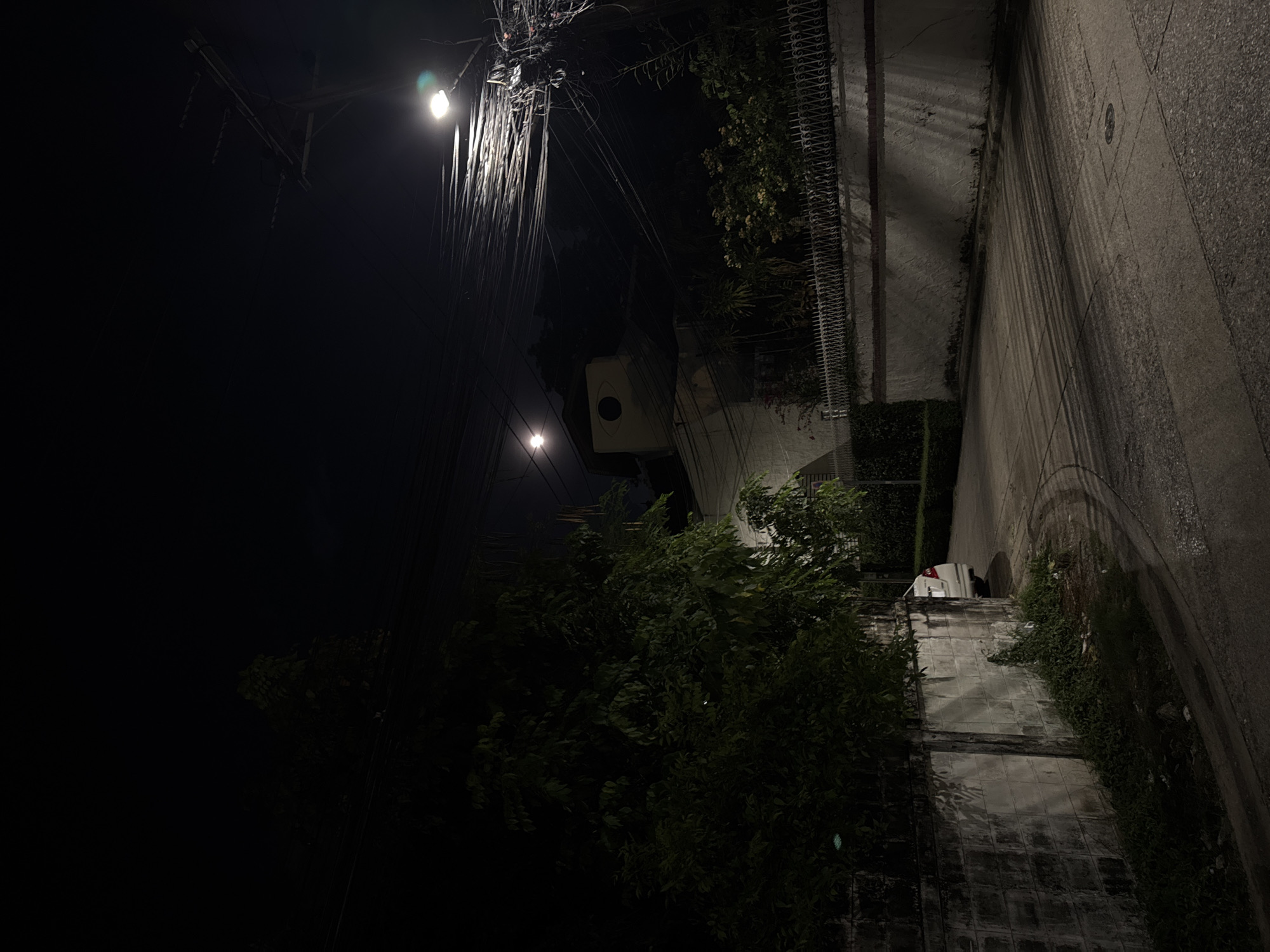 This nighttime photograph depicts a dimly lit urban street or alleyway, consistent with the provided context of Bangkok, Thailand. The scene is devoid of people or discernible activity, offering a quiet, static view.
The upper portion of the image is characterized by an extremely dense and tangled network of overhead utility wires, illuminated primarily by a bright streetlight in the upper left corner. A second, smaller streetlight is visible further down the left side. Beneath the wires, a light-colored, rectangular box-like structure with a dark circular opening is partially visible, potentially an electrical component or an exhaust vent.
Dense green vegetation, including bushes and small trees, occupies the mid-ground and lower-left sections, casting deep shadows. A decorative metal fence or railing runs along the top of a wall or building section, partially obscured by the foliage. The right side of the frame features a paved ground surface, likely a road or sidewalk, exhibiting textures and long, parallel shadows stretching across its surface, originating from the overhead wires and other unseen structures. In the lower right, behind a wall and some greenery, a portion of a white vehicle (possibly a truck or van) is visible.
The image is captured at night, with artificial street lighting creating strong contrasts and highlighting the complex web of wires and the textures of the ground and walls. No visible text is present. The chaotic arrangement of the utility lines is a notable detail, strongly suggestive of the specified location.