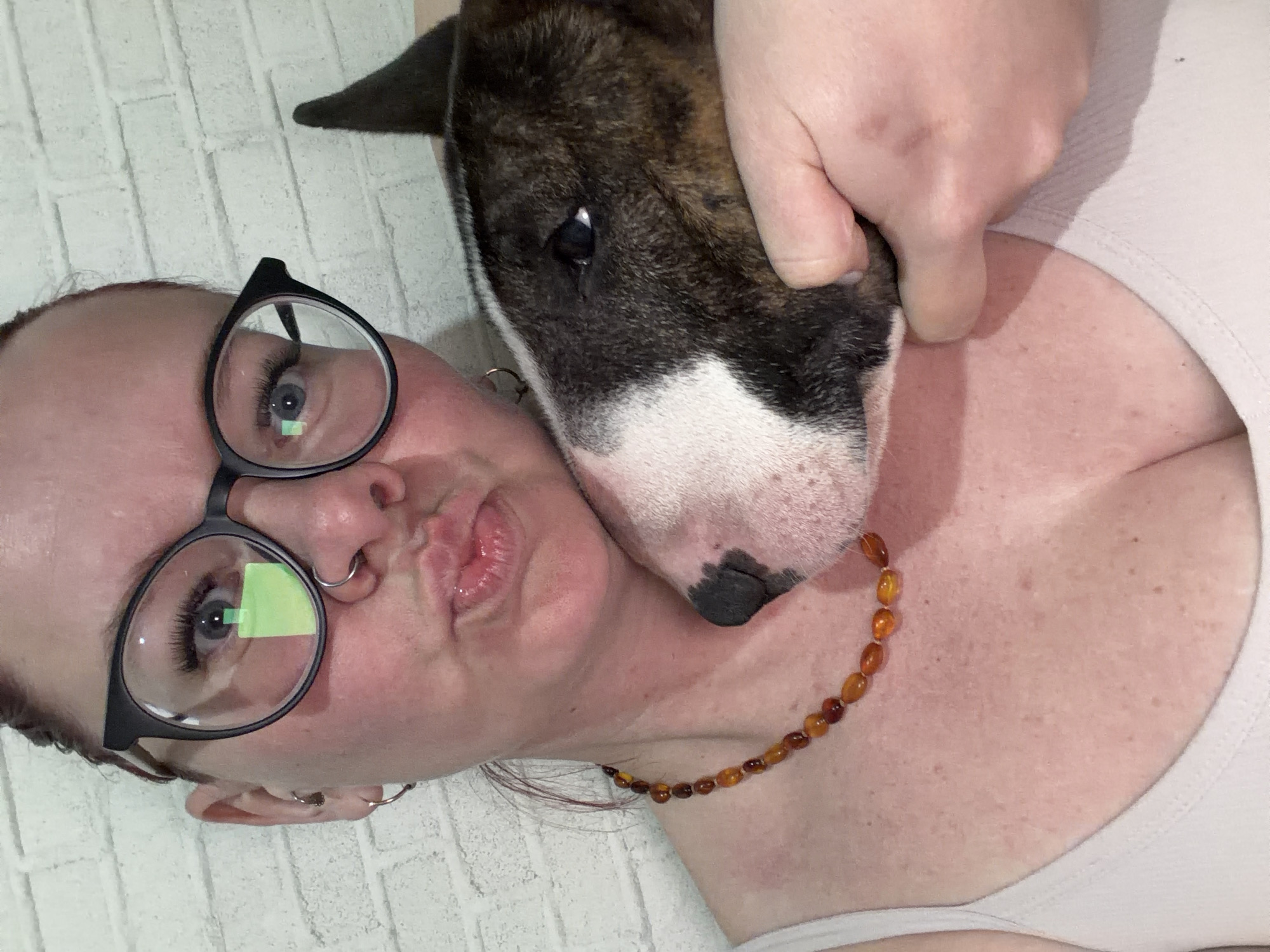 A woman with red hair and round black glasses is lying down next to a bull terrier. The woman is wearing a white tank top and an amber necklace. She has a nose ring and earrings. The bull terrier is mostly brown and white with a black nose. The location is not discernible from this image, and there is no evidence to indicate that it is Chaikovsky, Russia.