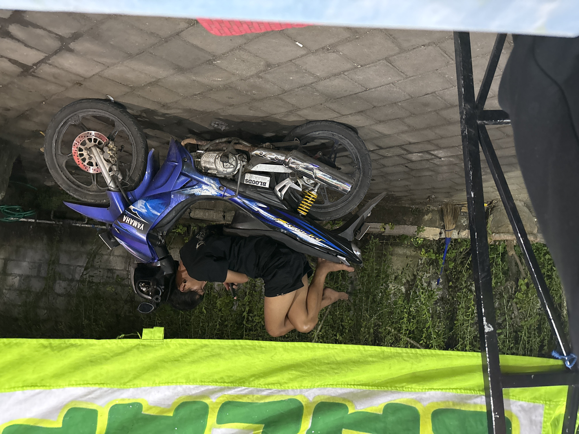 A young man with dark hair and earrings is lying on his back on a blue Yamaha motorcycle. He is wearing a black t-shirt and black shorts, and he is holding a smartphone in his hands, looking down at it. The motorcycle is parked on a paved area with brick-like stones. Behind the motorcycle, there is a wall covered in dense green foliage. A bright green banner with yellow lettering is stretched across the top of the frame, above the foliage. To the left of the motorcycle, a black metal structure is visible. A broom with a blue handle leans against the foliage near the motorcycle. The scene appears to be an outdoor area, possibly a parking spot or a roadside rest area, in Kuta Selatan, Indonesia.