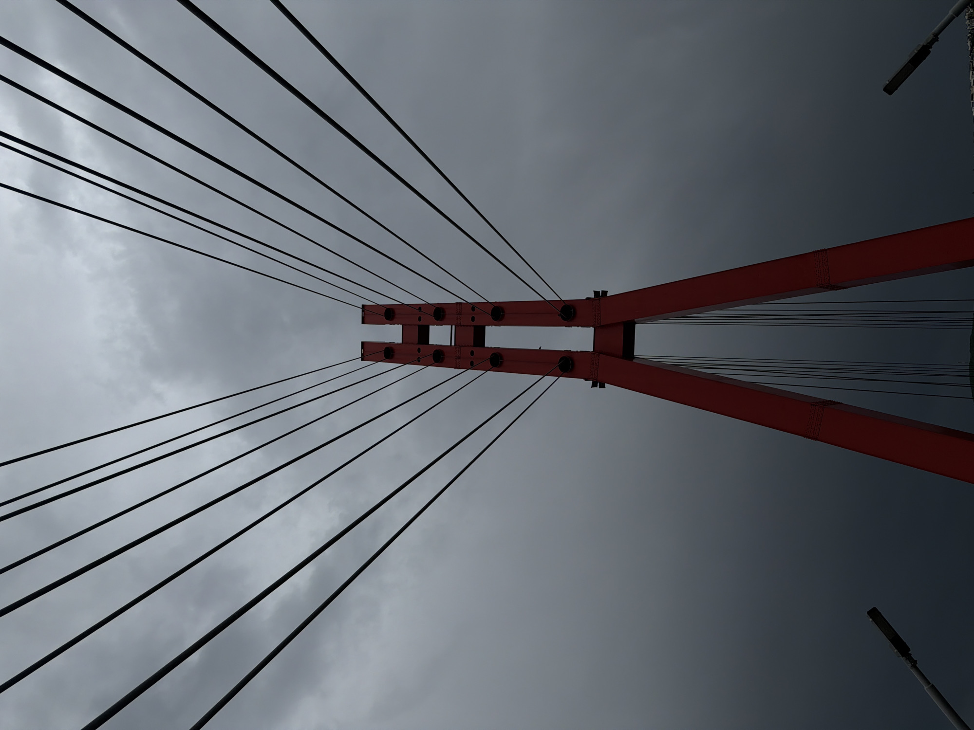 A low-angle shot looking directly upwards at a section of the Bridge of Peace in Tbilisi, Georgia, on a heavily overcast day. The primary focal point is a red structural element of the bridge, from which numerous black suspension cables extend upwards and outwards. The background is a uniform, dark grey sky filled with thick clouds. A few lampposts are visible in the top right and bottom right corners. The overall composition highlights the architectural and engineering design of the bridge against the somber sky.