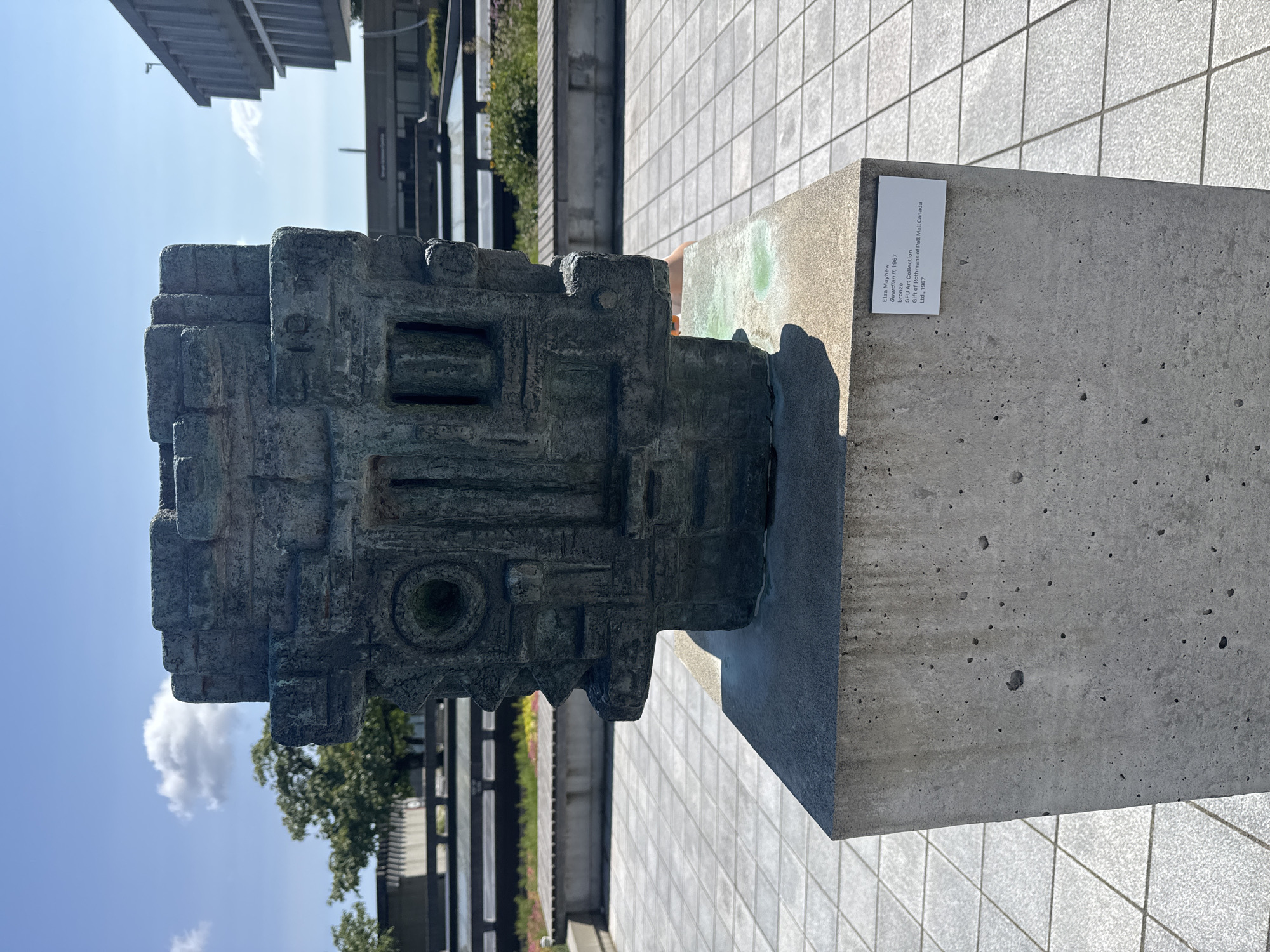 An outdoor, close-up shot captures a weathered bronze sculpture titled "Guardian II," created by artist Elsa Mayhew in 1967. The sculpture, displayed on a concrete pedestal, features an abstract, geometric form with multiple recessed areas and a circular opening. A plaque on the pedestal identifies the artwork, noting it is part of the SFU Art Collection and was a gift from Rothmans of Pall Mall Canada, Ltd. in 1967. The background reveals a modern architectural setting with buildings featuring large windows and concrete elements, characteristic of the Simon Fraser University campus in Burnaby, Canada. The ground is paved with square tiles, and benches with planters filled with colorful flowers are visible in the middle ground. The sky is blue with a few white clouds, suggesting a clear day. No people are clearly visible in the frame.