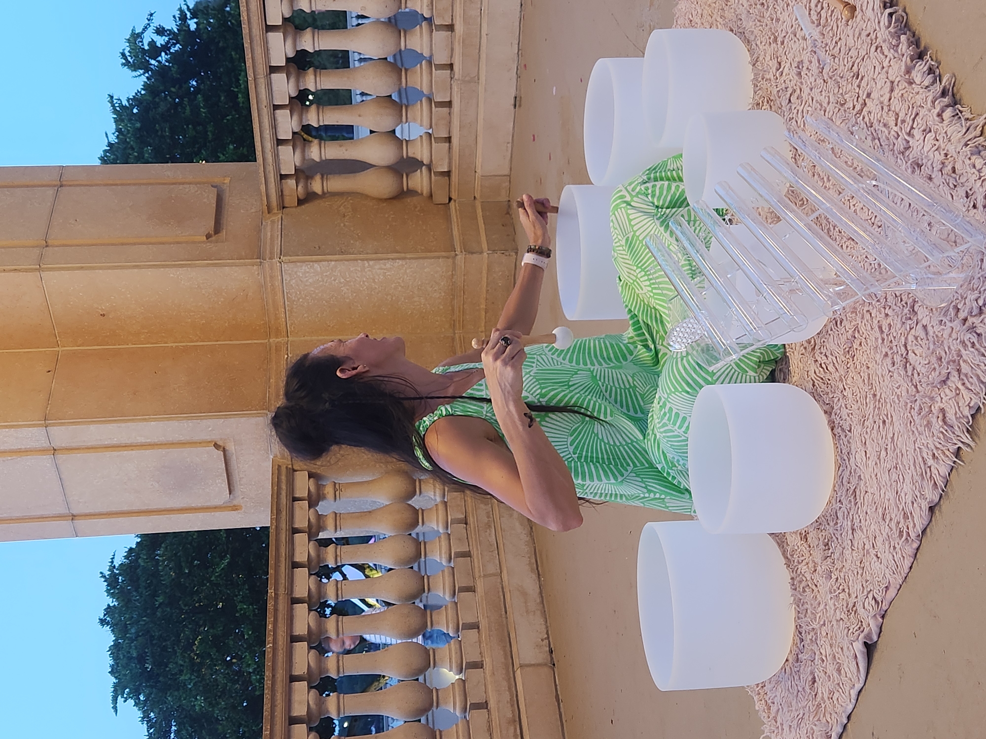A woman in a green and white patterned dress is kneeling on a pink rug in front of several large, frosted white bowls. She is holding a wooden mallet with a white rubber tip in her right hand and is about to strike one of the bowls. Her left hand is also near the bowls. The bowls appear to be crystal singing bowls, and a clear acrylic stand with multiple grooves is visible in front of them. Behind the woman, a stone balustrade with ornate pillars separates the foreground from a background with trees under a twilight sky. The scene is set outdoors on what appears to be a covered porch or balcony of a building with light-colored stone architecture. There is a faint bokeh effect on the background, suggesting it is a shallow depth of field. The woman has dark hair pulled up into a bun, and a small butterfly tattoo is visible on her left arm. A child, out of focus, can be seen through the balustrade in the mid-ground. The location is Altamonte Springs, United States of America.