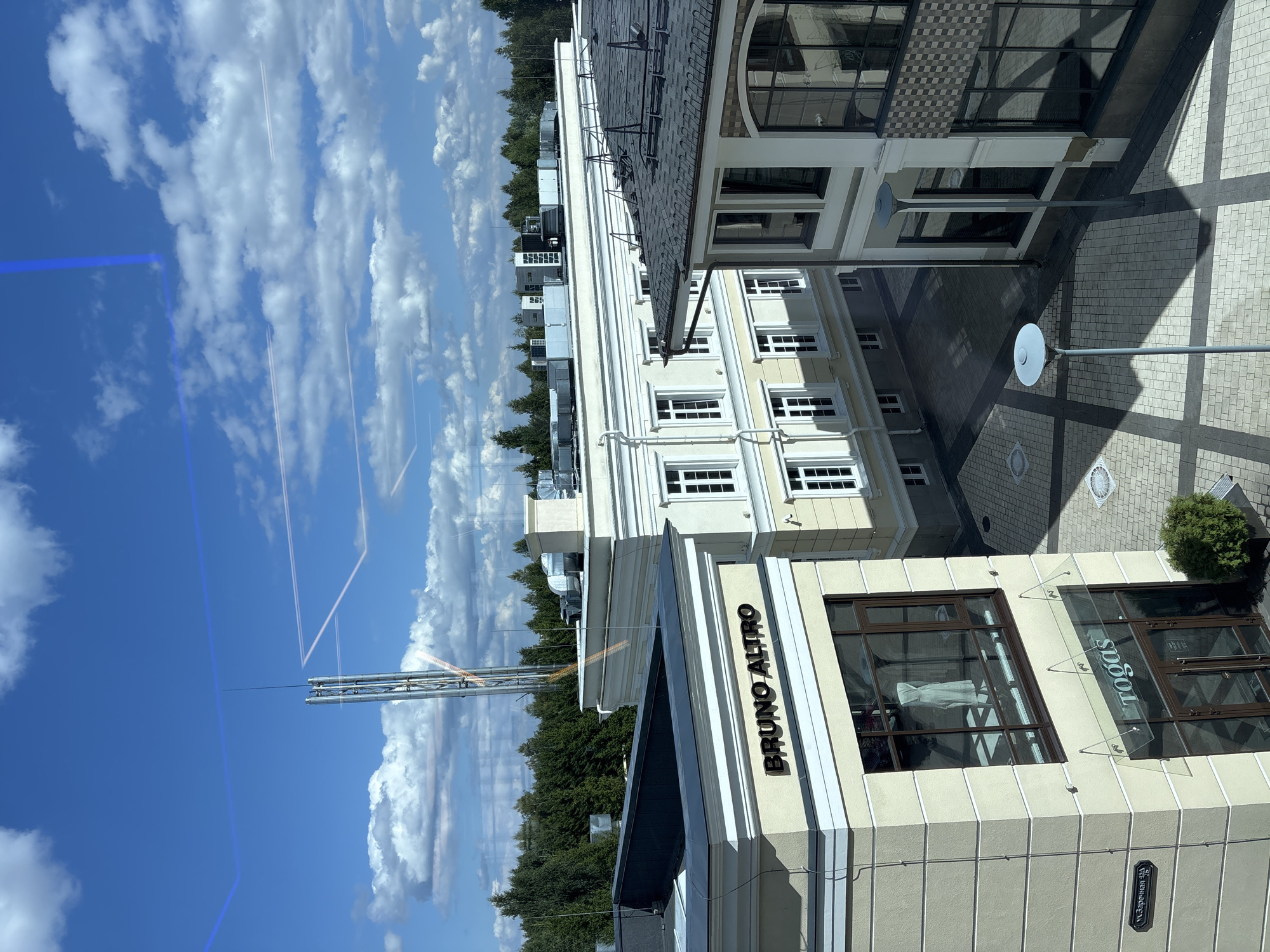 The image is a high-angle shot of a modern urban complex on a sunny day, with a bright blue sky and scattered white clouds. The architecture features light-colored buildings with multiple windows and dark tiled roofs. Some buildings have classical elements like decorative cornices. One building has a sign that reads "BRUNO ALTRO" and another below it reads "TOGAS". The complex is surrounded by a dense forest. In the distance, a tall, industrial-looking tower or structure is visible against the sky. The ground area between the buildings is paved with interlocking stones, and there are streetlights and some greenery. Reflections of blue lines and a white rectangle are visible on the glass surface from which the photo was taken, indicating it is likely taken from an interior looking out. The location is identified as Monolit, Russia. There are no people visible in the image.