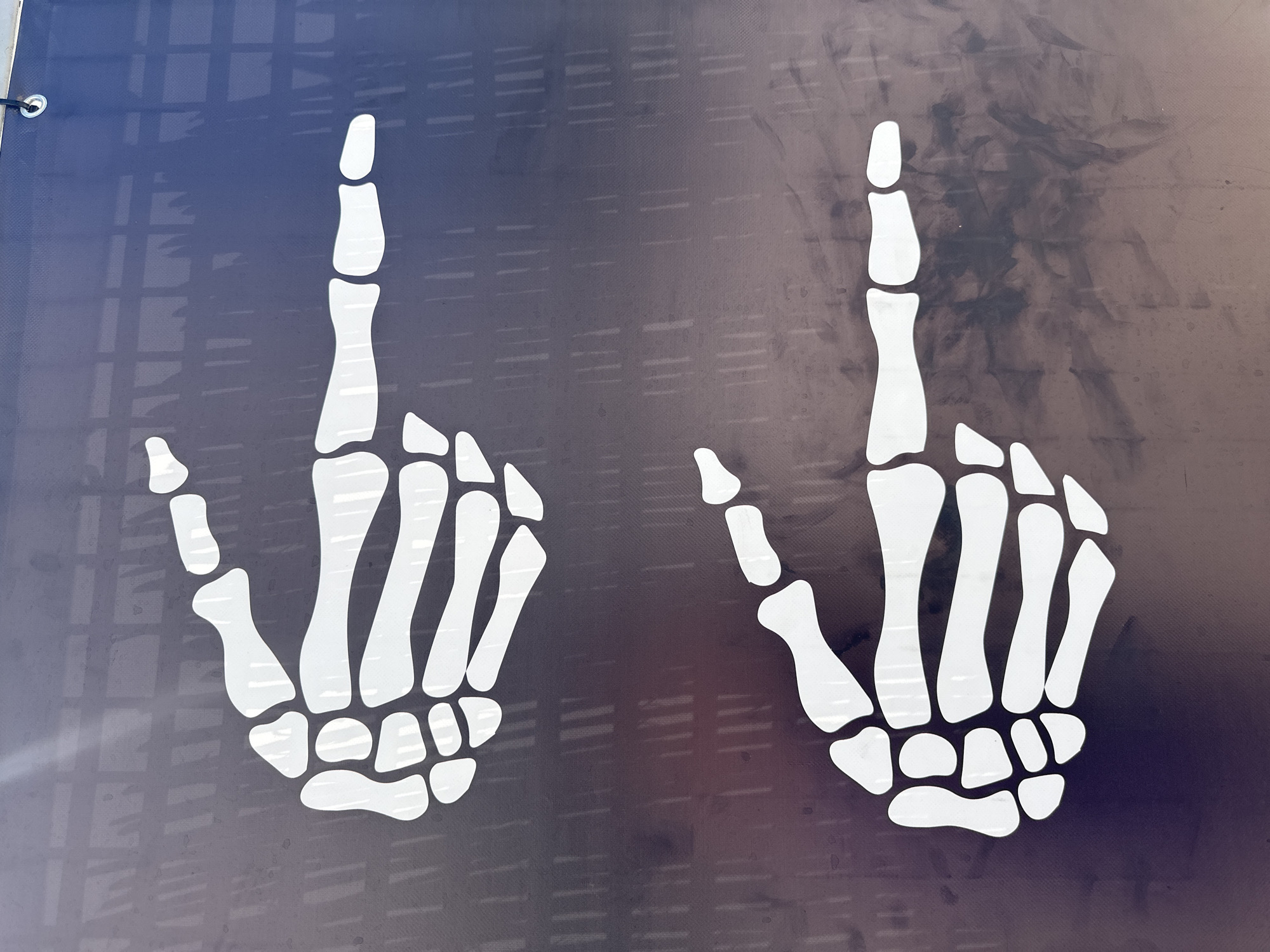 This image shows two white skeletal hand designs against a dark blue-grey background. Both hands are posed with only the index finger extended upwards, resembling the number one. The background is slightly textured with some vertical lines. The image was likely taken outdoors, given the natural lighting and the presence of a metal grommet in the upper left corner. The location, Eivissa, Spain, cannot be verified from this image.