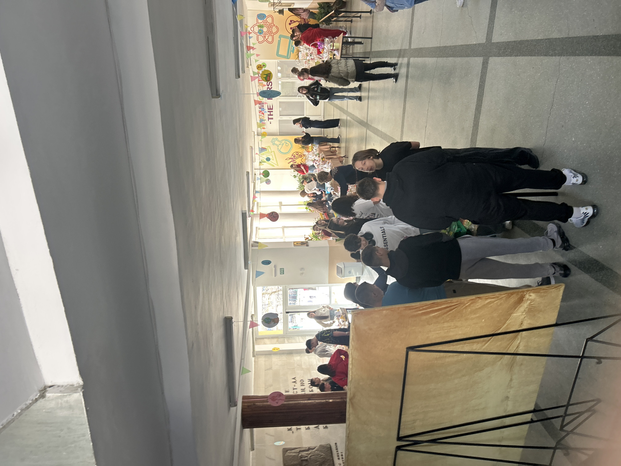 The image captures a crowded hallway scene in Zlatograd, Bulgaria, possibly during an event. The space is decorated with colorful garlands, balloons, and wall decorations, suggesting a festive or celebratory occasion. Numerous people are present, including groups of students and adults, some standing and others gathered around tables or displays. A table is covered in gold cloth. The hallway features a tiled floor with dark lines and a high ceiling with visible vents. The environment suggests an indoor location, likely a school or community center, due to the presence of educational wall displays.