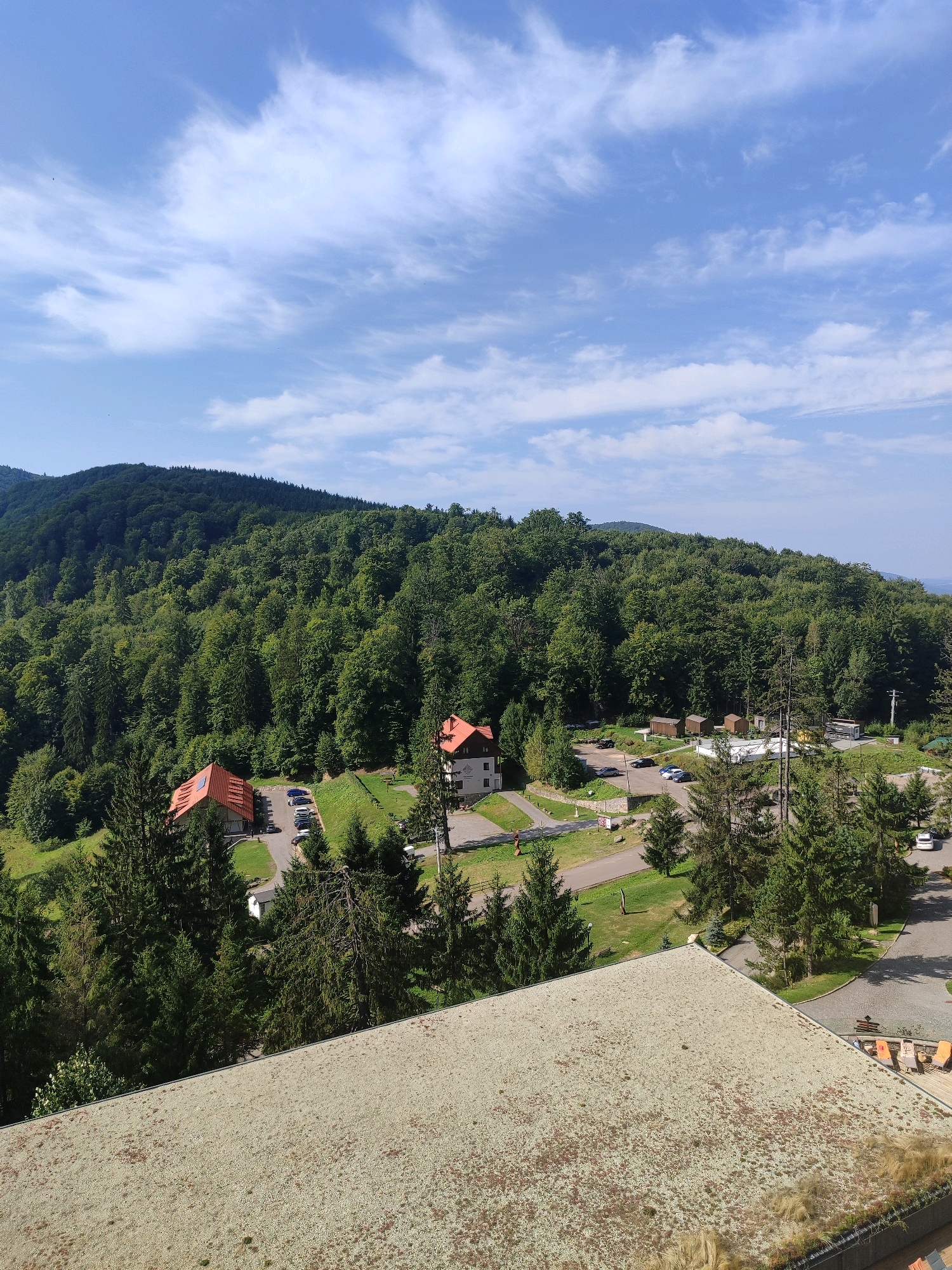 The image is a wide shot of a scenic landscape in Băile Bálványos, Romania, on a clear, sunny day with a partly cloudy sky. In the foreground, a large, weathered, light-green roof covered in patches of moss and dirt occupies the lower portion of the frame. The midground features a dense, verdant forest covering rolling hills and mountains. Scattered within the treeline and on clearings are several buildings, including a white building with a red-tiled roof and another similar building. A paved road winds through the area, with several cars parked along its sides, suggesting a resort or residential area nestled within the natural landscape. Pine trees are interspersed throughout the scene, adding to the woodland atmosphere. The background is dominated by the expansive forest and the distant, hazy outline of mountains under a blue sky with scattered white clouds. The overall impression is one of a tranquil, mountainous retreat surrounded by nature.
