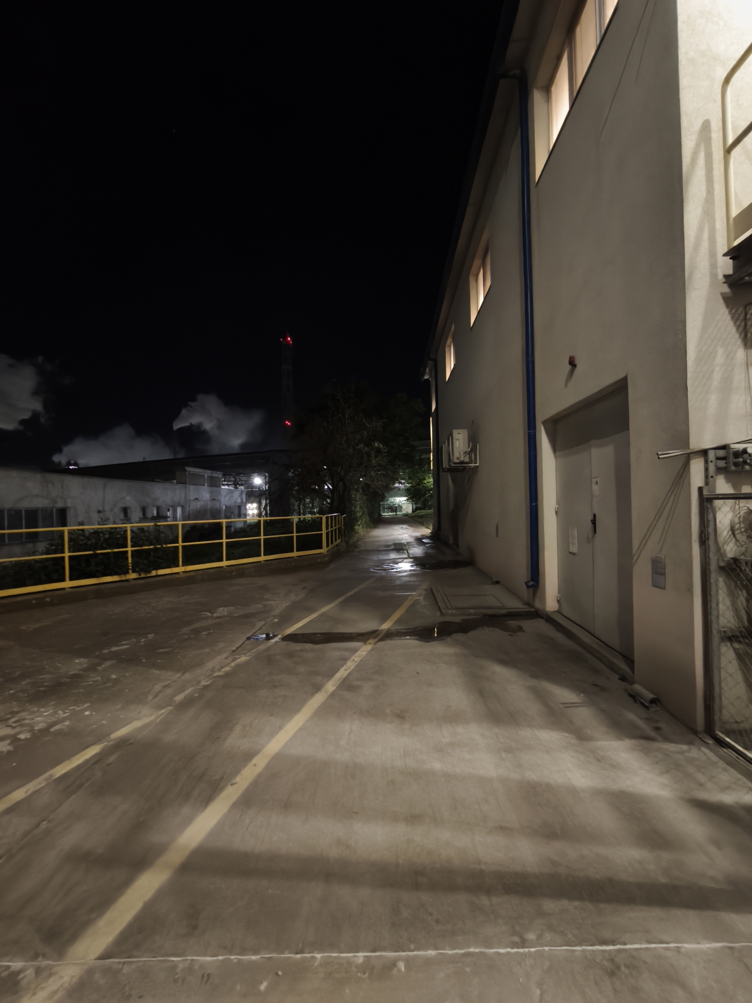 At night, the scene captures an industrial or warehouse area. A long, paved path stretches into the distance, flanked by a building on the right and a yellow guardrail on the left. In the distance, another building emits steam or smoke, with a red-lit tower rising above it. The night sky is dark, with minimal lighting illuminating the surroundings. The scene appears deserted, with no visible human activity.
