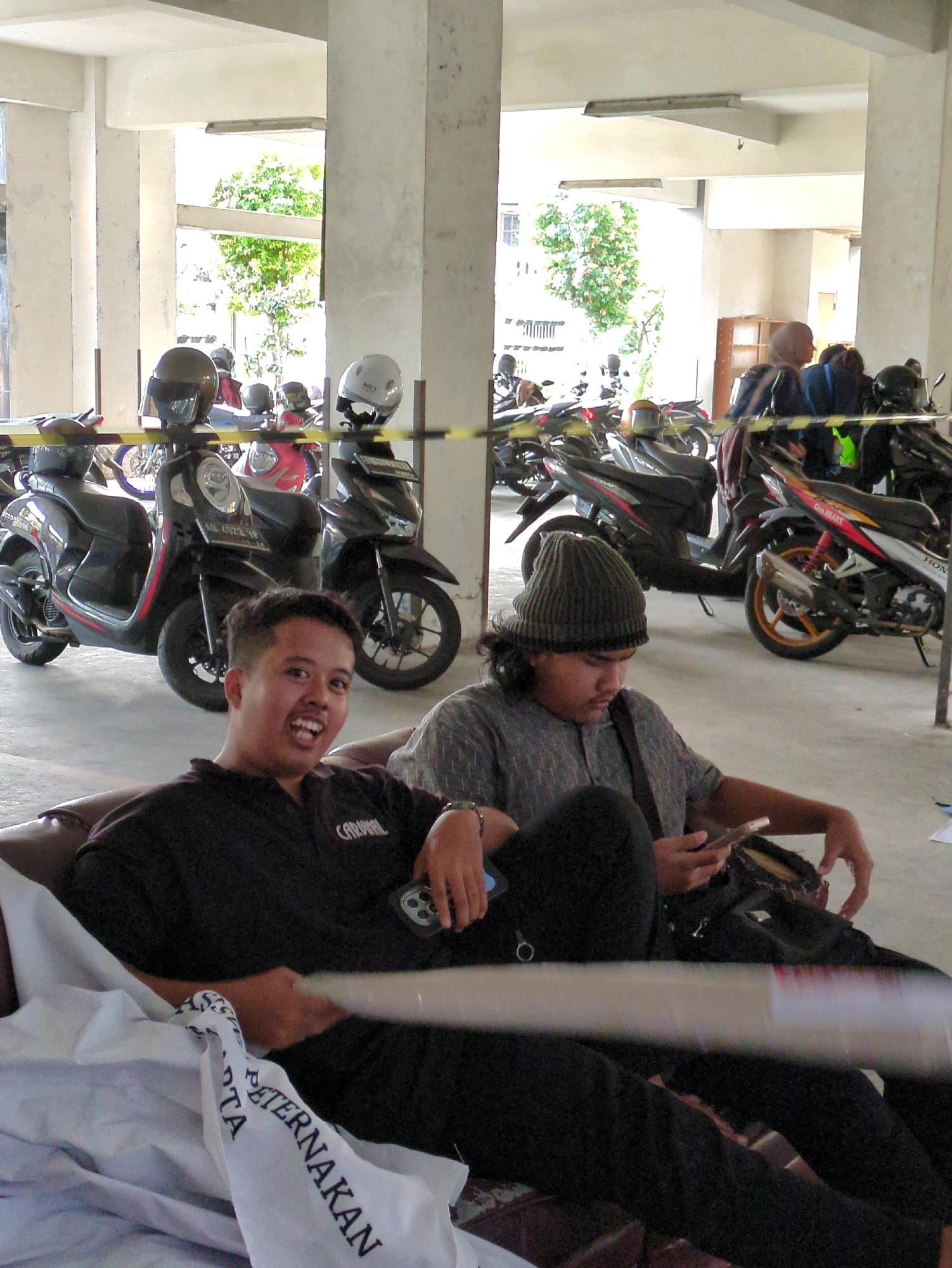 In Surakarta, Indonesia, two young men are seated on a brown couch in a covered outdoor parking area. The man on the left, facing the camera, has short dark hair and is wearing a black collared shirt with the word "CARDINAL" visible on it. He is smiling broadly and holding a smartphone in his right hand. To his right, the second man is wearing a grey beanie and a grey patterned shirt. He is looking down at his phone, which he holds in his left hand. Several motorcycles are parked behind them, and the area appears to be part of a building with concrete pillars and an open-air ceiling. A white cloth with the Indonesian word "PETERNAKAN" (livestock) printed on it is draped over the couch and the legs of the man on the left.